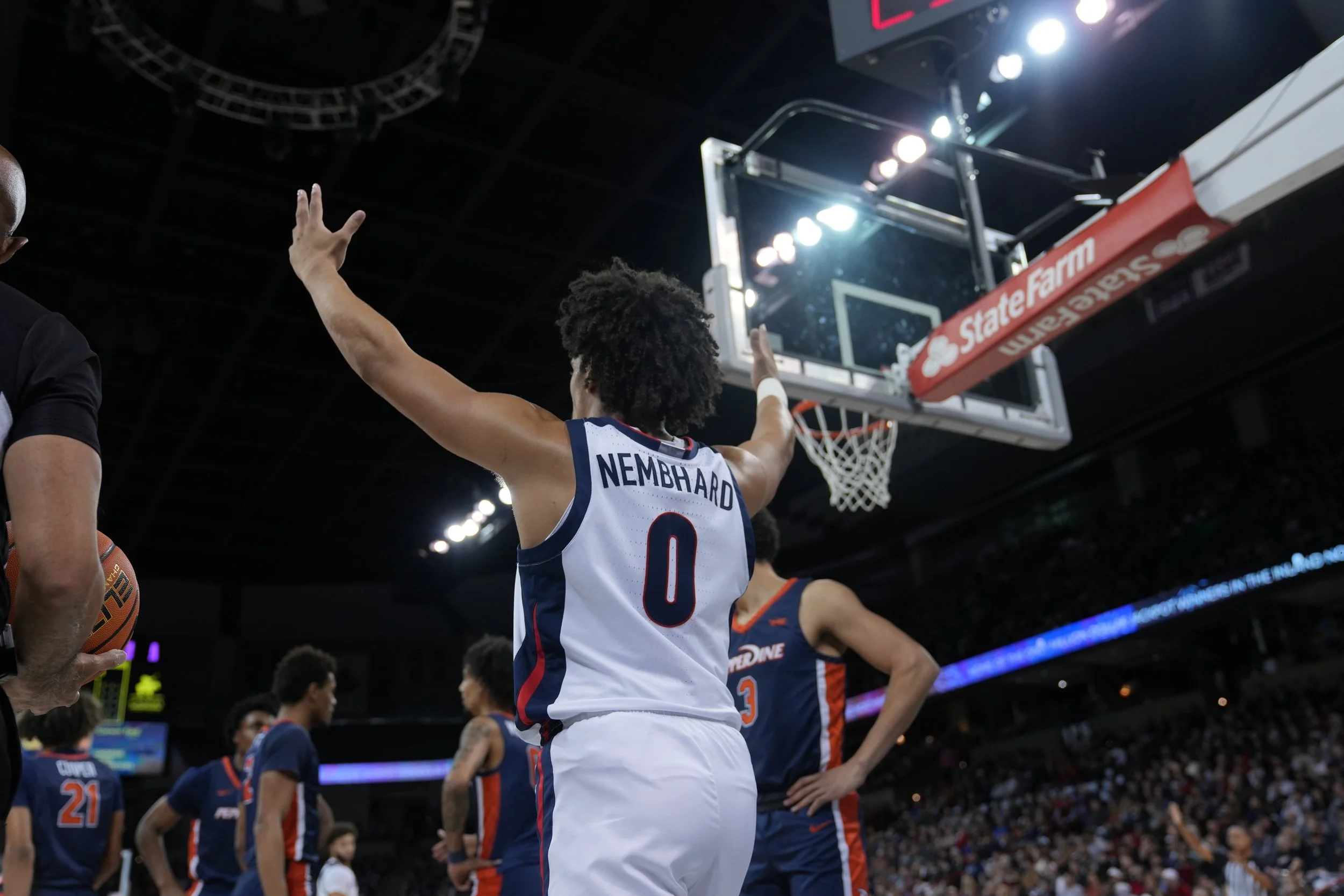 Basketball player in a white jersey with 'Nembhard 0' on the back raising arms near a basketball hoop during a game, with several players and a referee visible.