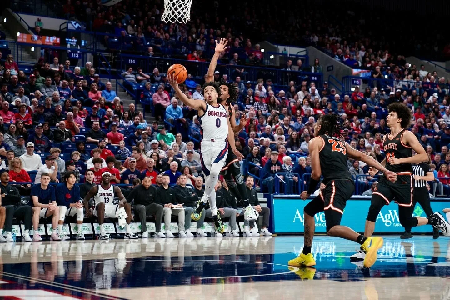 Basketball player in white uniform attempting a layup during a game, with opposing players in black uniforms defending, and a crowd watching from the stands.
