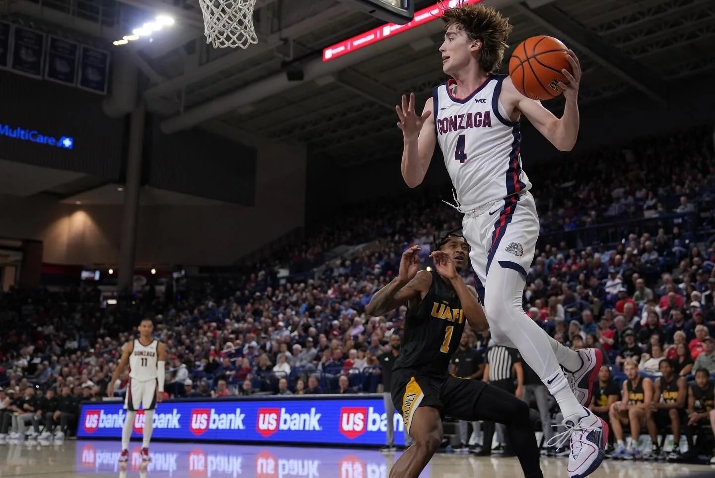 Basketball player in mid-air holding a ball near the hoop, with an opposing player nearby in a packed sports arena.