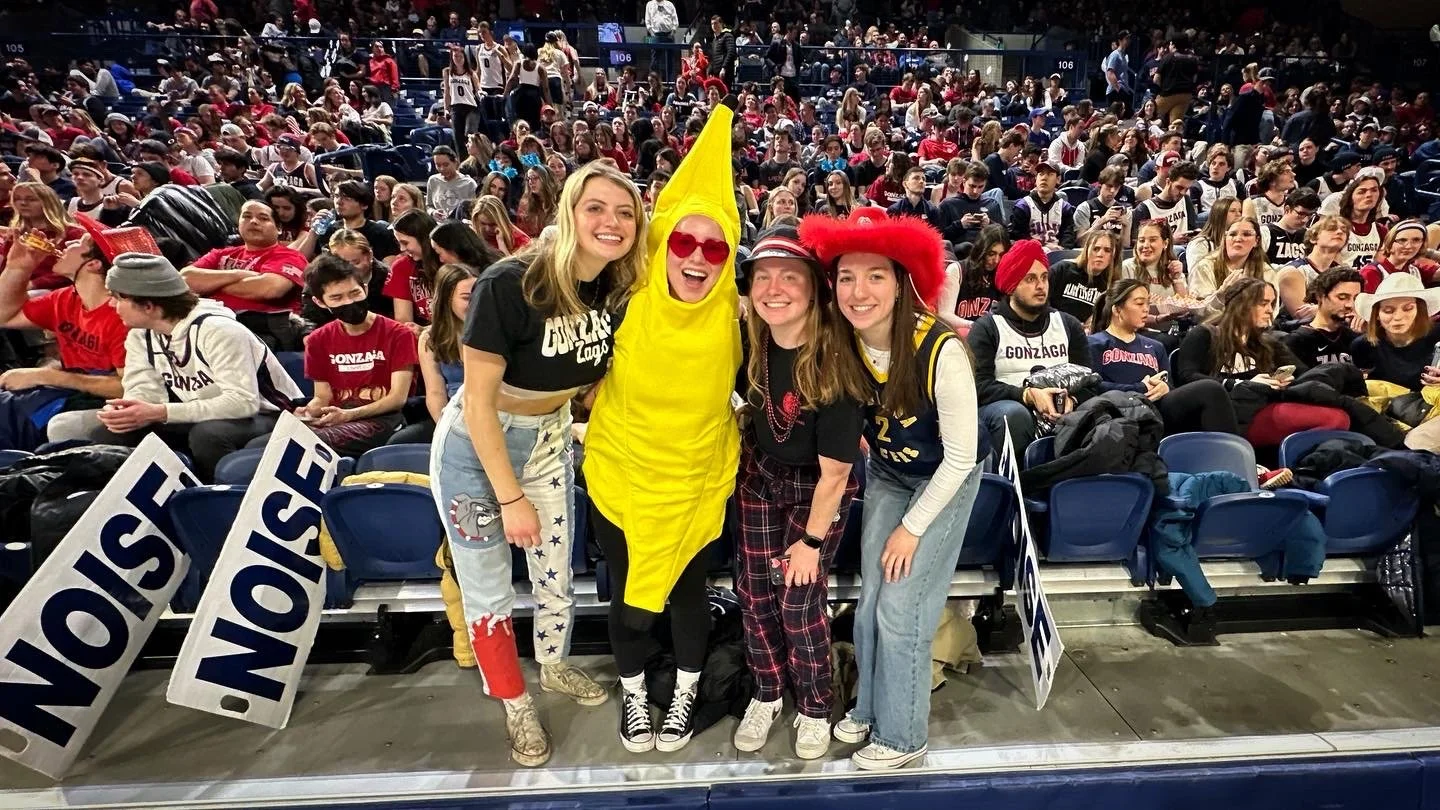 Group of fans in a stadium, one in a banana costume, others in casual attire, holding noise signs. Large cheering crowd in the background.