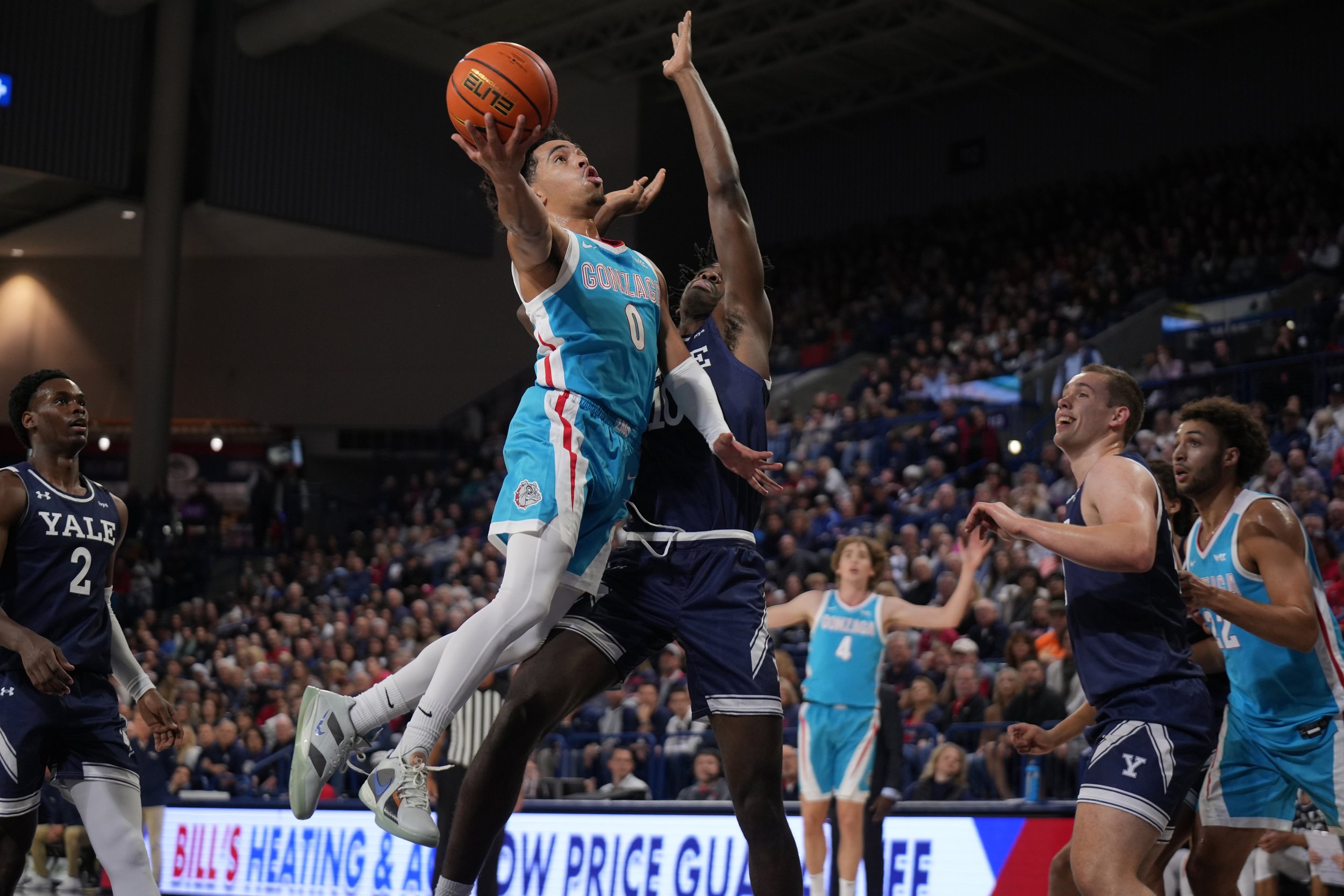 Basketball game with players from two teams; a player in a blue uniform jumps with the ball towards the hoop while being defended by players in dark blue uniforms. Spectators are visible in the background.