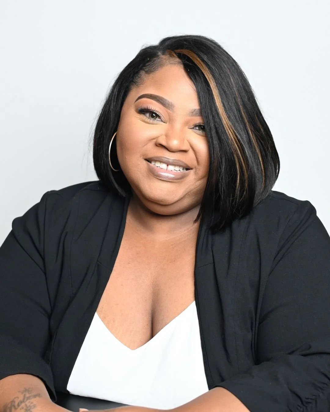 A woman with shoulder-length black hair with blonde highlights, wearing a black blazer over a white top, and gold hoop earrings, smiling against a plain light gray background.