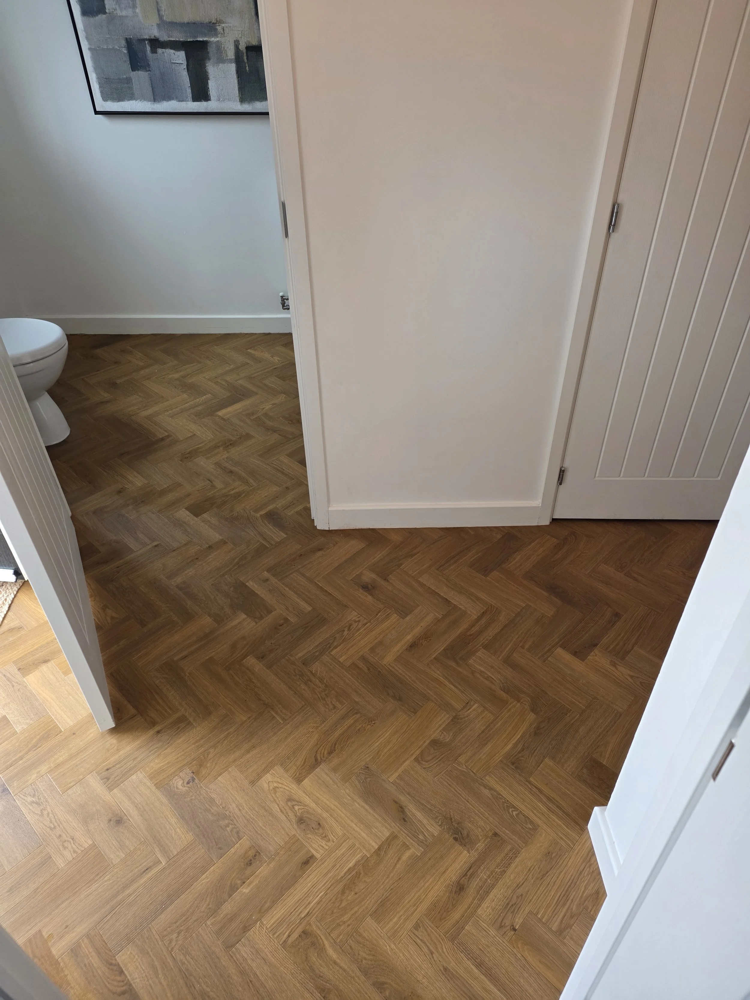 View of a room with wooden parquet flooring, white walls, an abstract art piece on the wall, and part of a white door on the right side. A restroom is partially visible through an open door.
