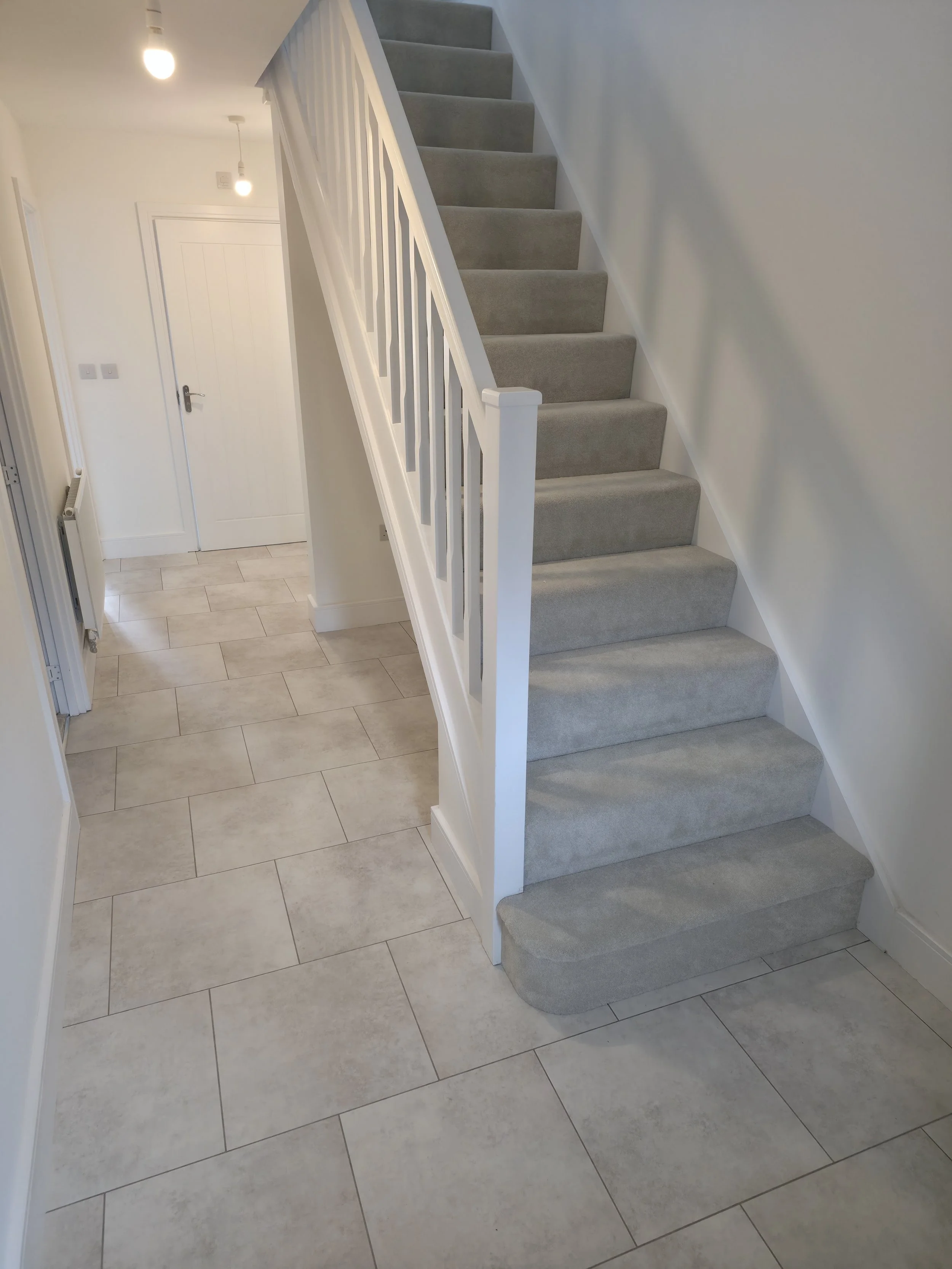 Interior view of a modern home with a carpeted staircase, white walls, tiled flooring, and ceiling lights.