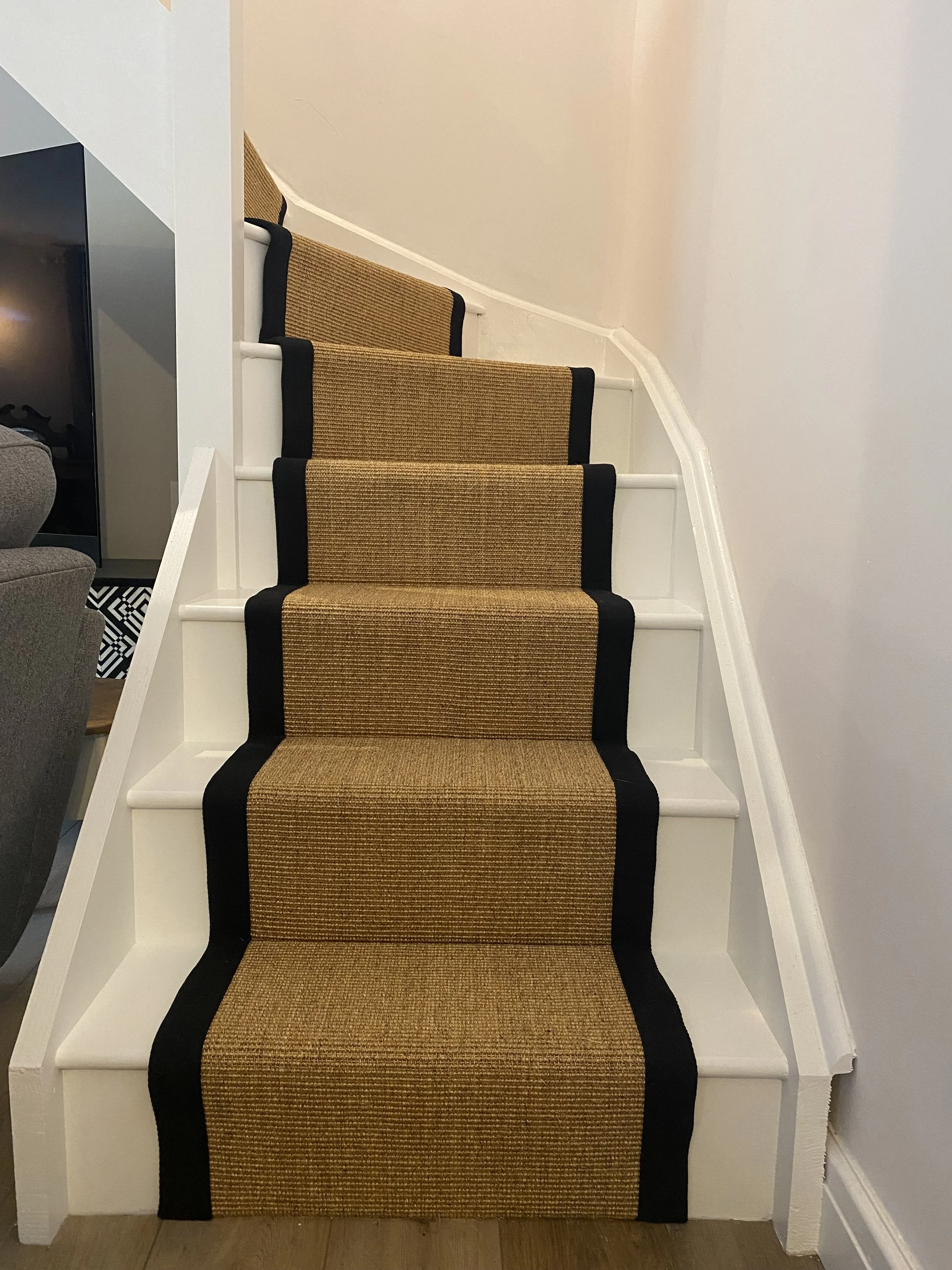 Interior staircase with a beige and black runner rug, white stairs, and a white wall with a curved molding on the right side.
