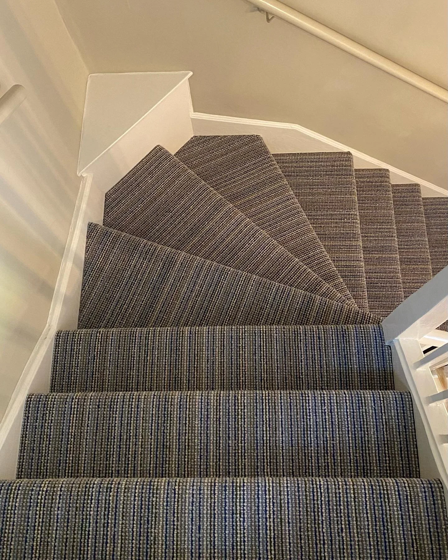 A view looking down a staircase with multi-colored striped carpet, white walls, and a white handrail.
