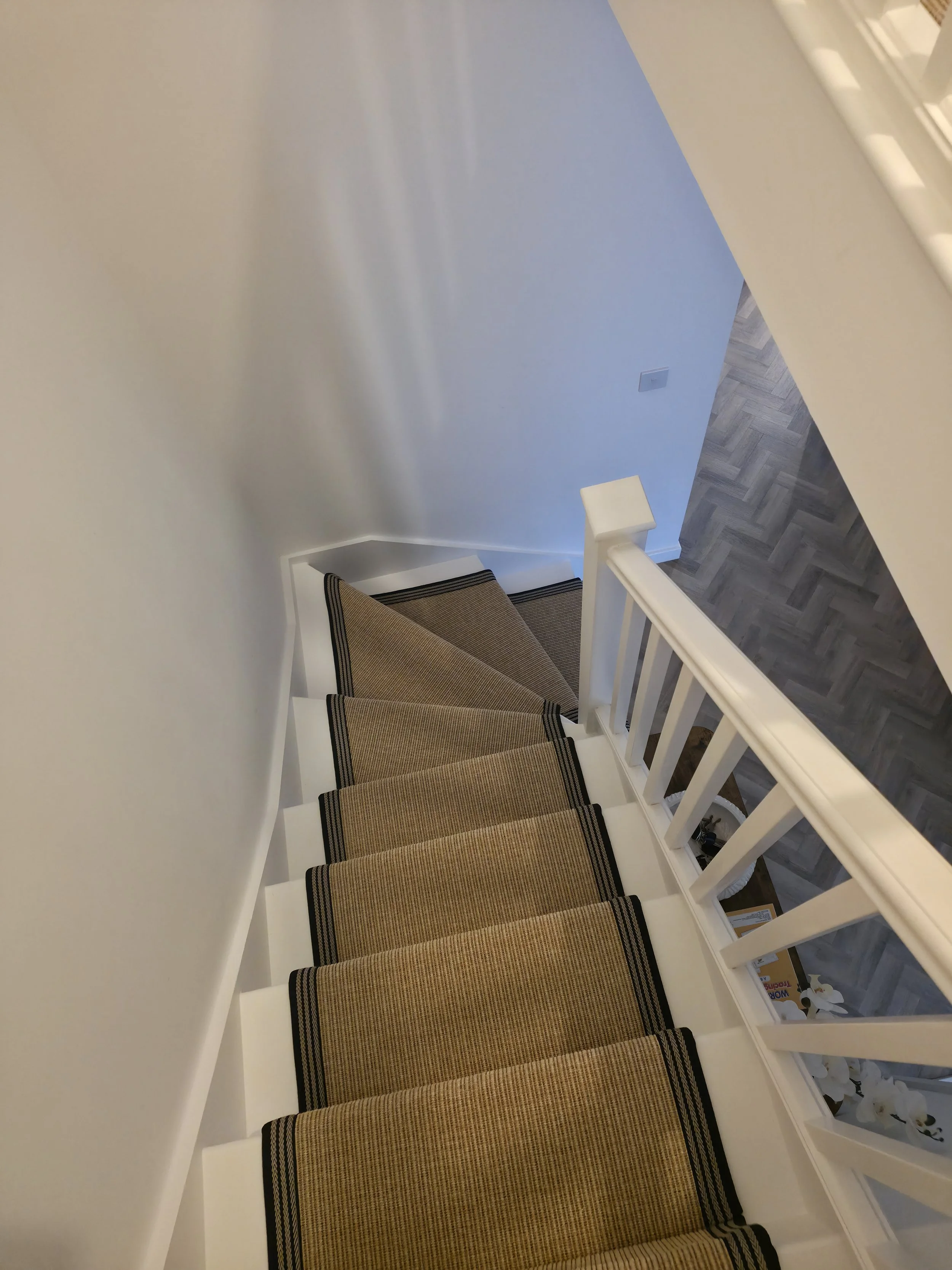A staircase with beige carpeted steps and a black border, alongside a white railing, leading down to a landing with grey patterned flooring, next to a white wall with an electrical outlet and a blue-painted wall.