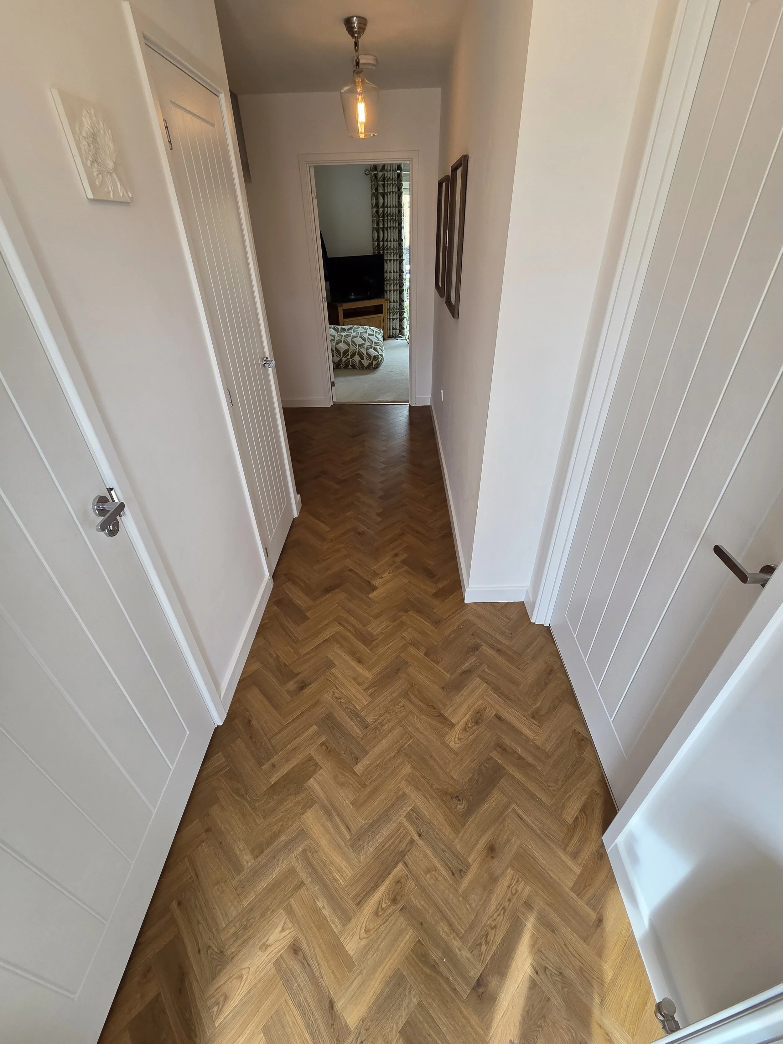 View of a hallway with white-paneled closet doors on either side, leading to a room with a television, a patterned ottoman, and patterned curtains, a wood floor in a herringbone style, and a ceiling light fixture.
