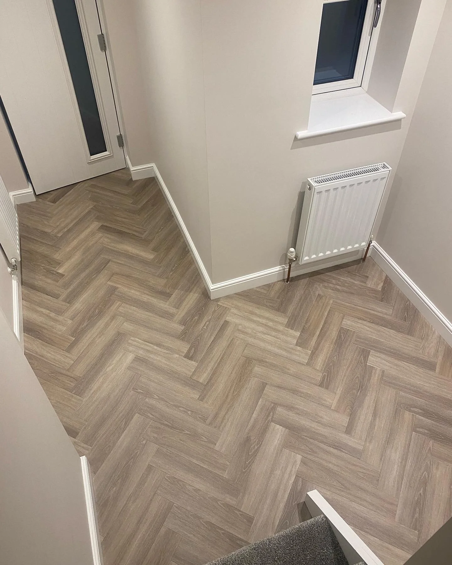 A corner of a room with beige herringbone patterned wooden flooring, a white radiator, a window with a white sill, and a partially visible white door with a vertical glass panel.