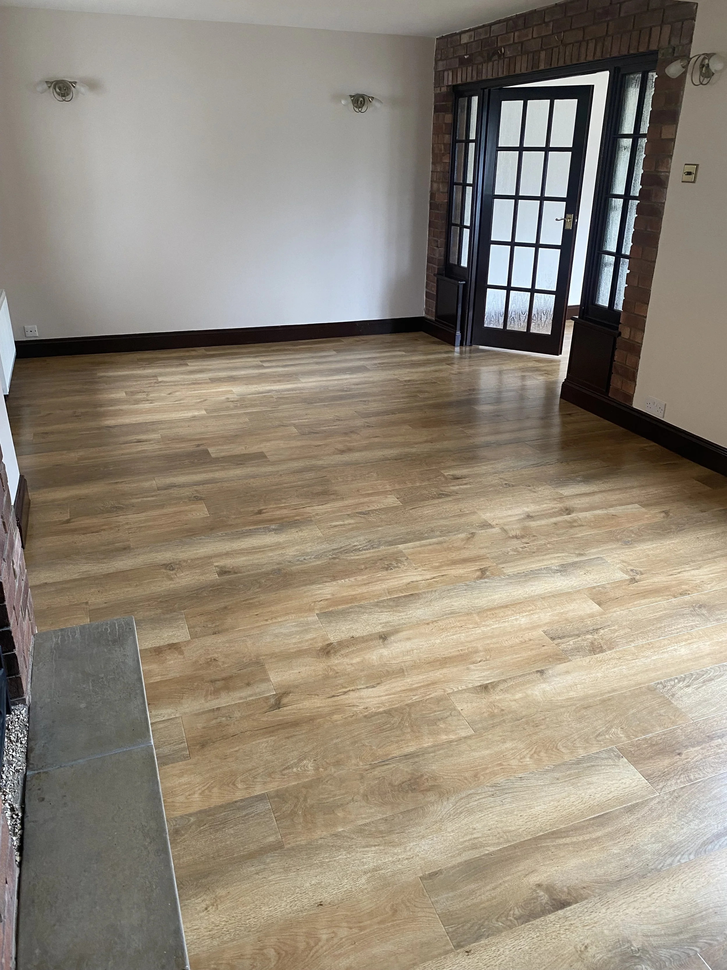 Empty room with wooden flooring, white walls, wall-mounted lights, and a large glass door with black framing surrounded by exposed red brick.
