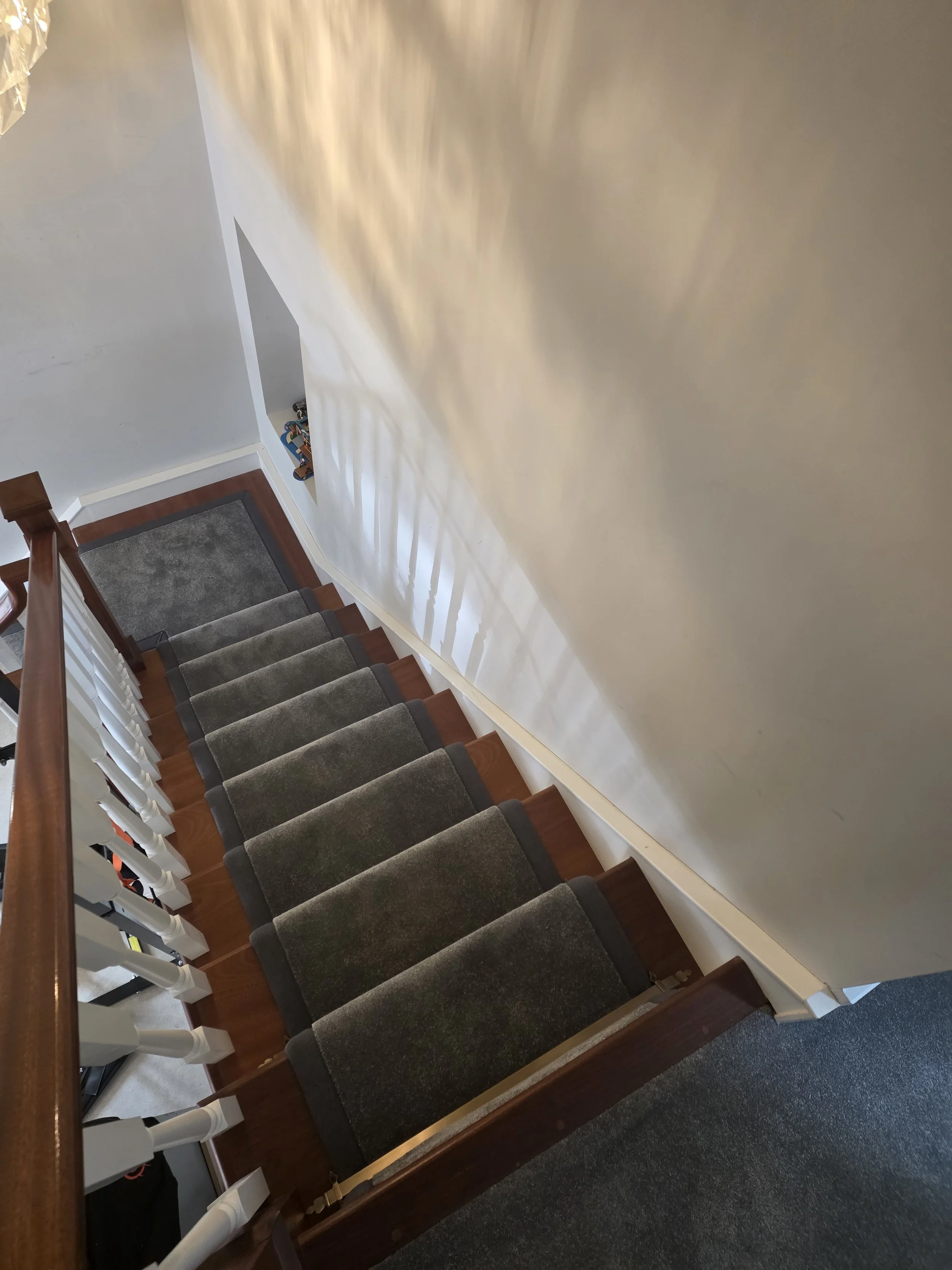 Winding staircase with gray carpet runner, wooden handrail, and shadow patterns on a beige wall.