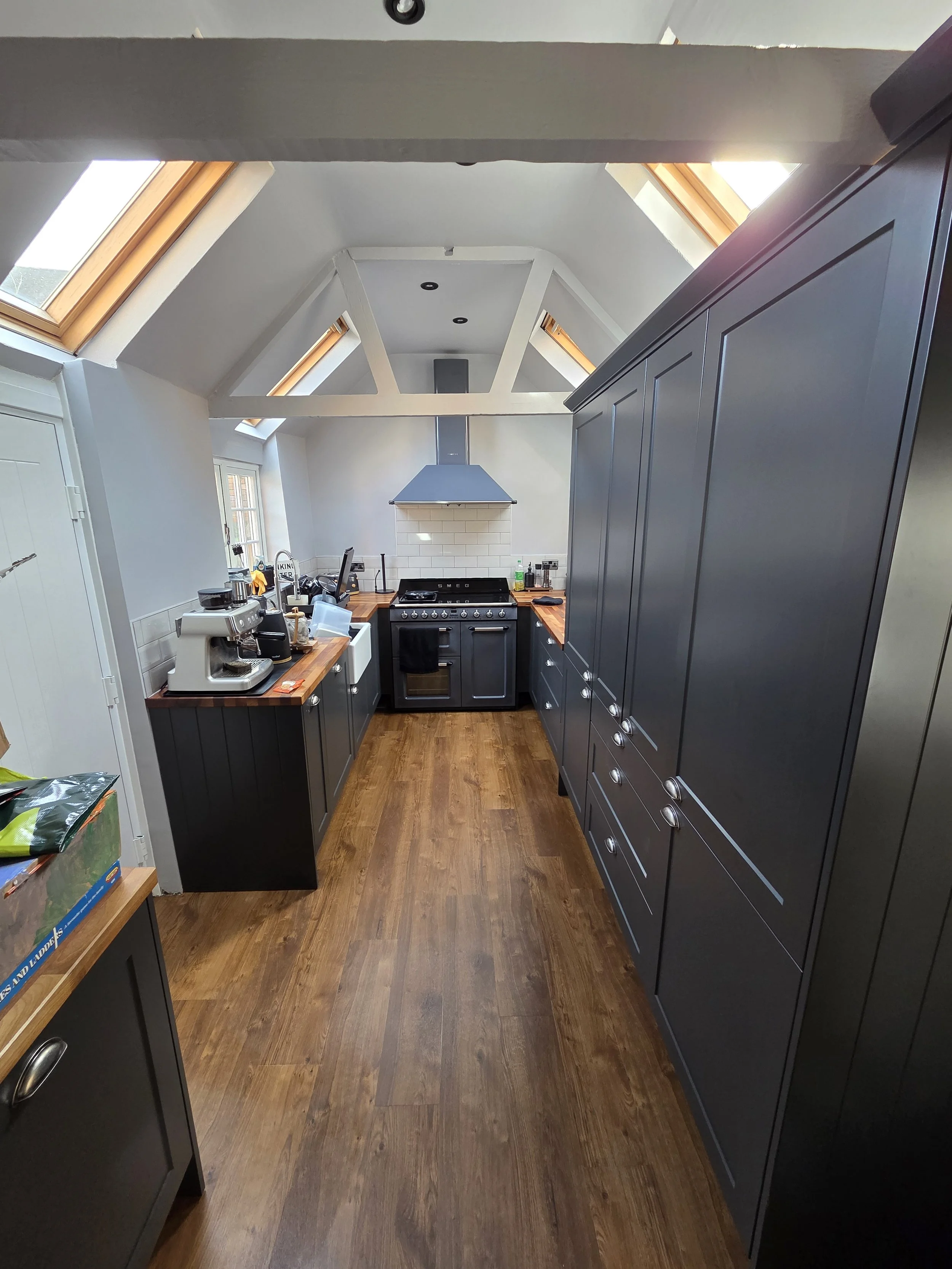 A small kitchen with a sloped ceiling, skylights, and wooden flooring. Blue cabinets line one side, and a countertop with kitchen appliances and items is visible. The stove is positioned at the end of the kitchen with a chimney above.