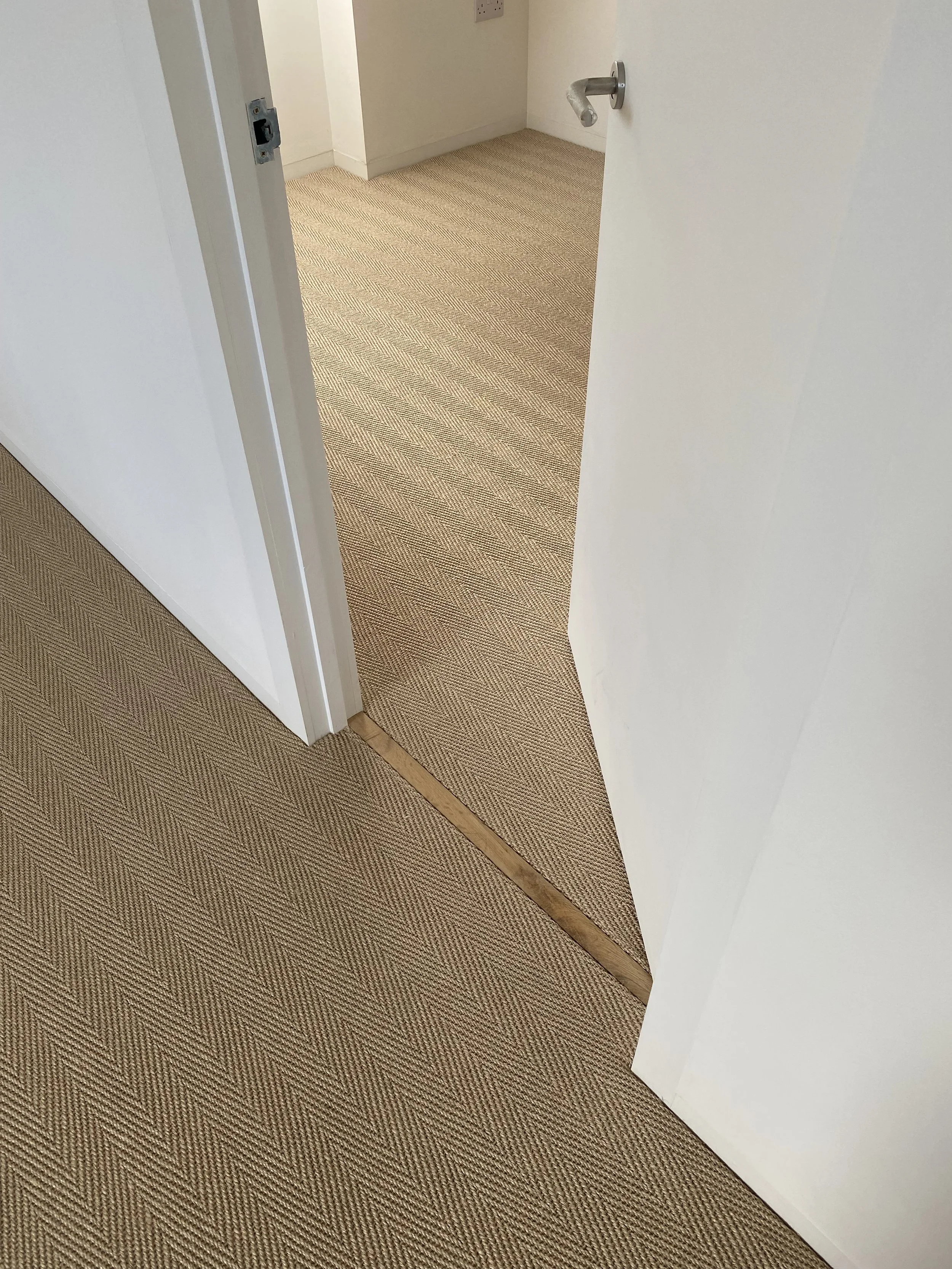 An open white door with a metallic handle leads to a room with a patterned beige and brown carpet flooring and a white wall.