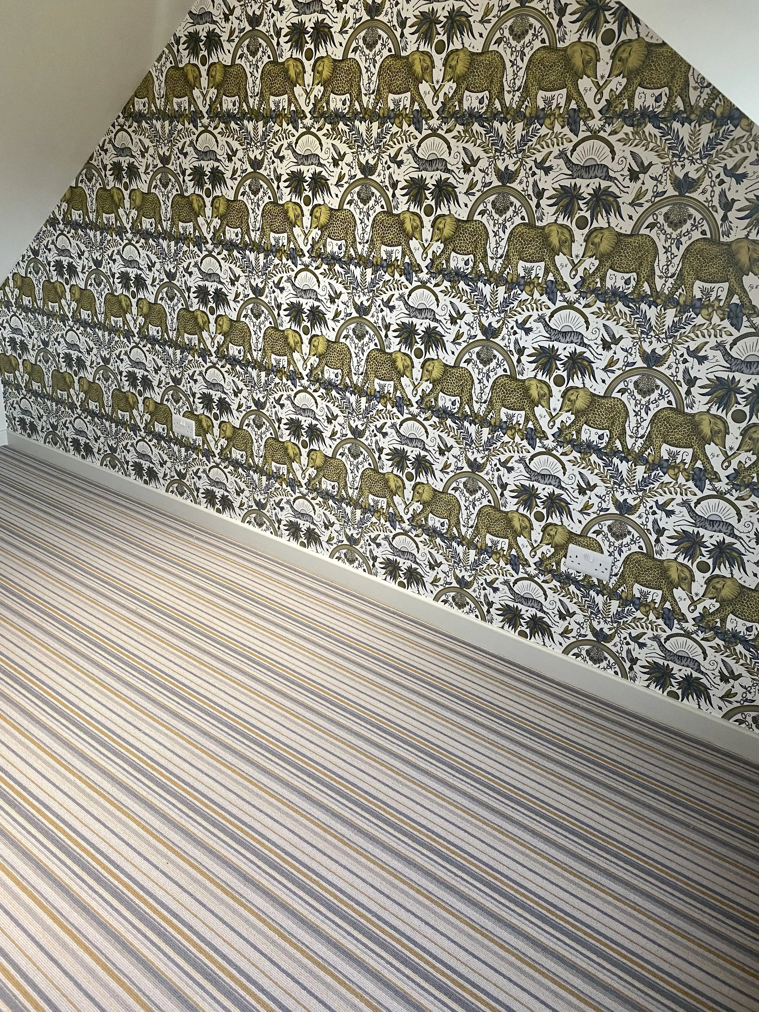 Decorative wallpaper with a pattern of elephants, trees, and foliage in shades of green and black, on a white background. The wall has a sloped ceiling and a striped carpet in neutral tones.