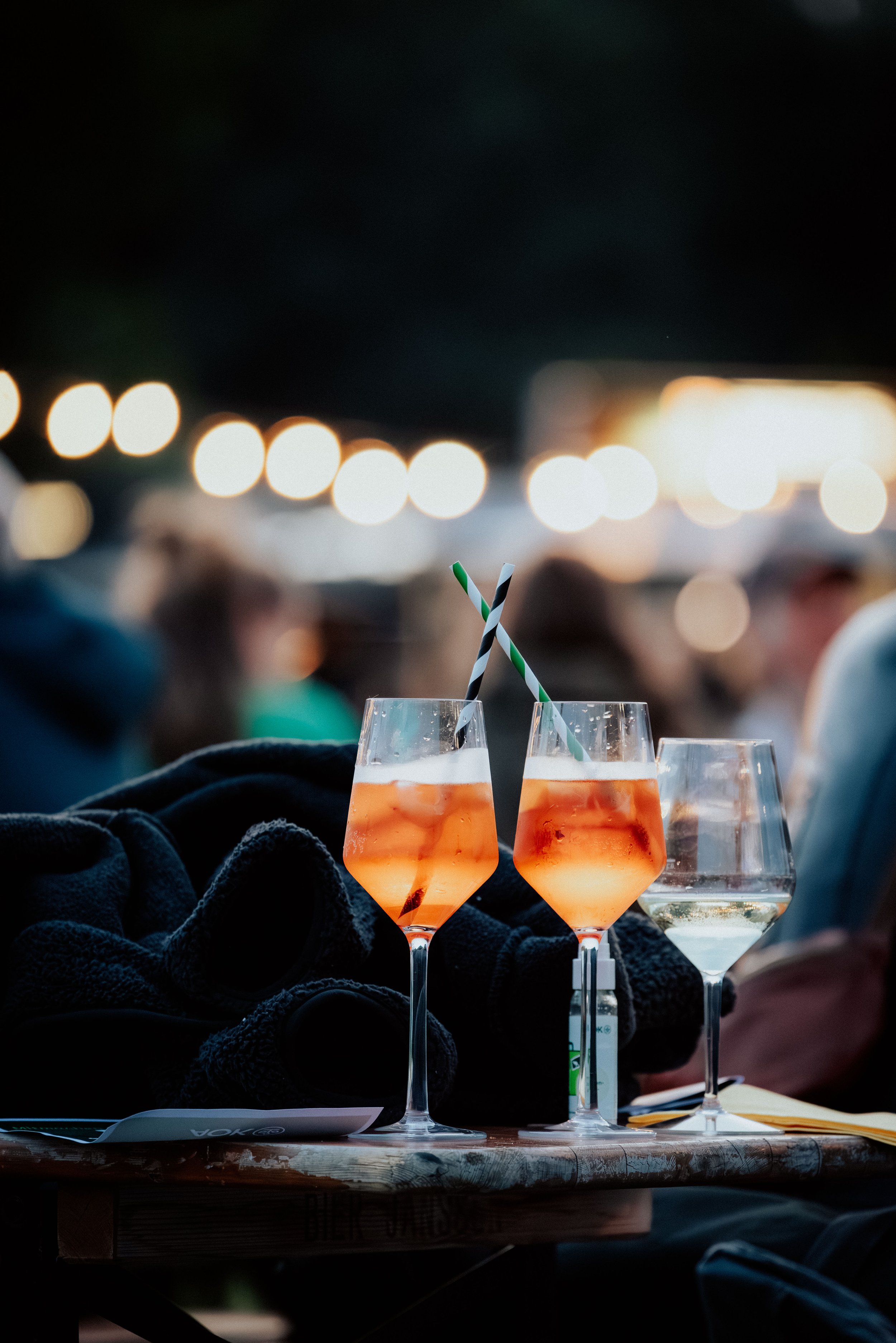 Zwei Gläser mit orangefarbenem Getränk und Strohhalmen auf einem Tisch bei einer Feier im Freien bei Nacht, im Hintergrund unscharfe Lichter und Menschen.