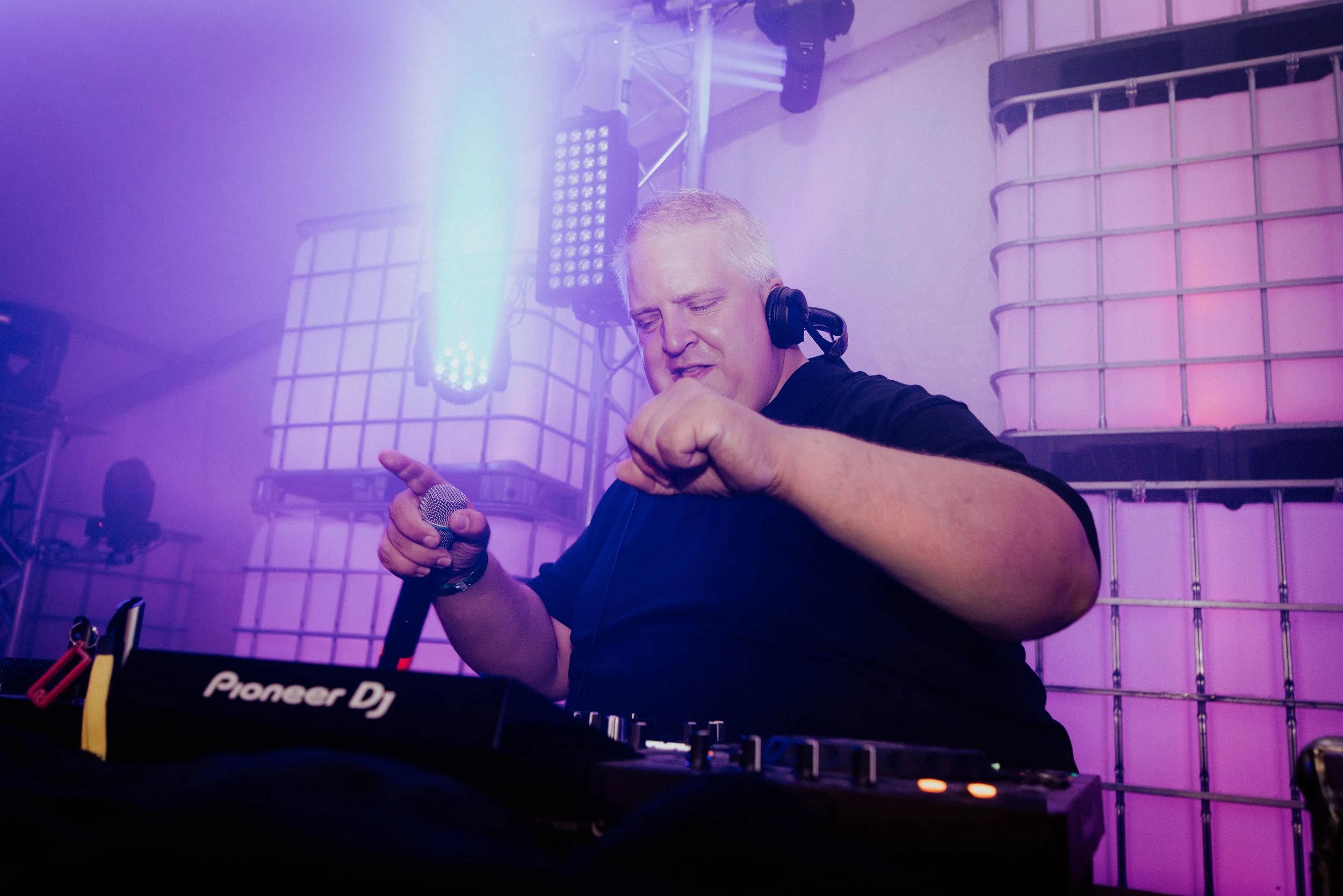 Ein DJ mit kurzen grauen Haaren, der mit Kopfhörern auf dem Kopf Musik macht, während er an einem Mischpult bei einer Party oder einem Event arbeitet.