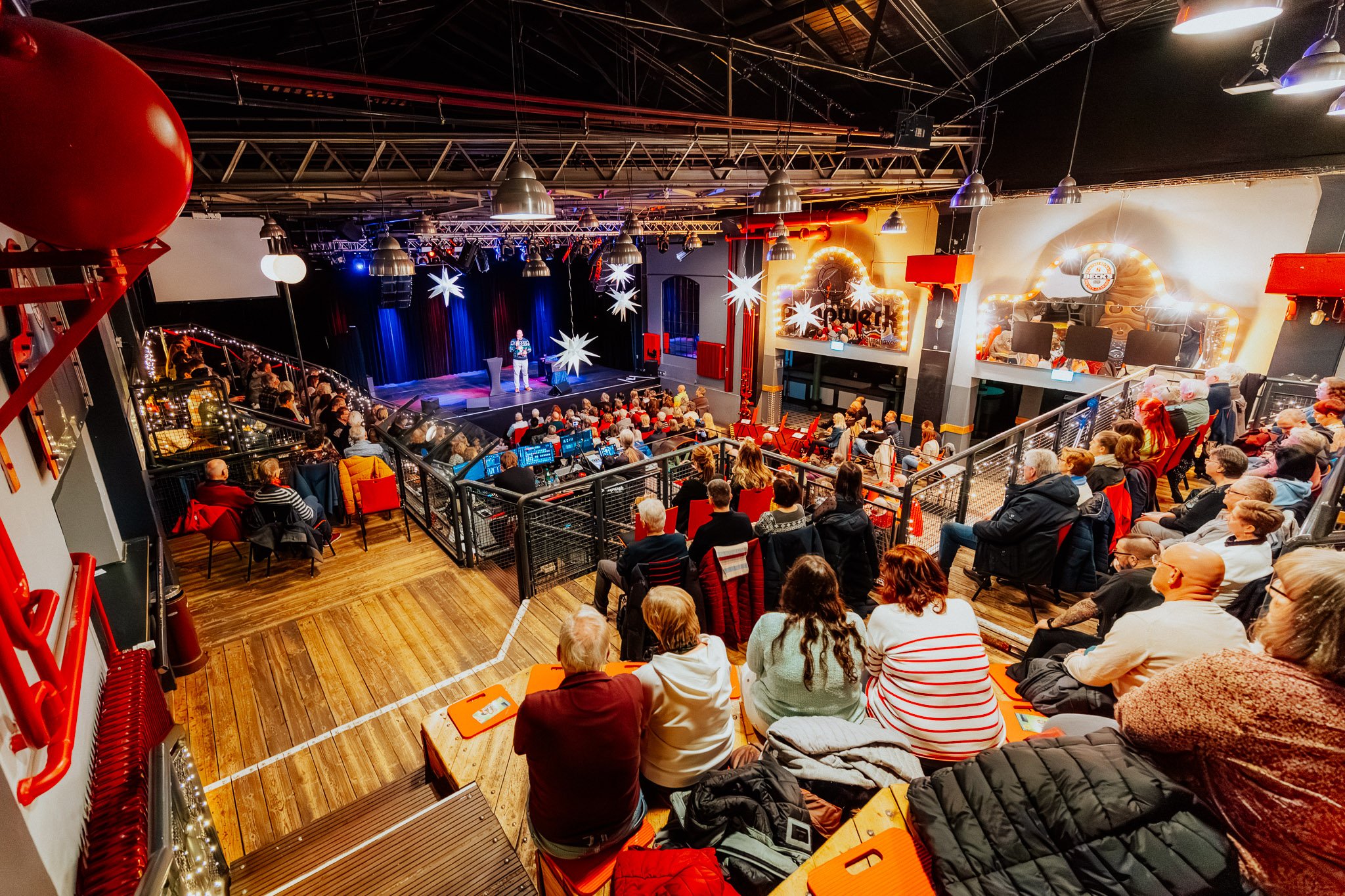 Auf einer Bühne in einem Veranstaltungsraum steht ein Mann beim Comedy- oder Kabarett-Programm, die Bühne ist mit weißen Sternen dekoriert, das Publikum sitzt in mehreren Reihen und schaut zu. Der Raum ist festlich beleuchtet.