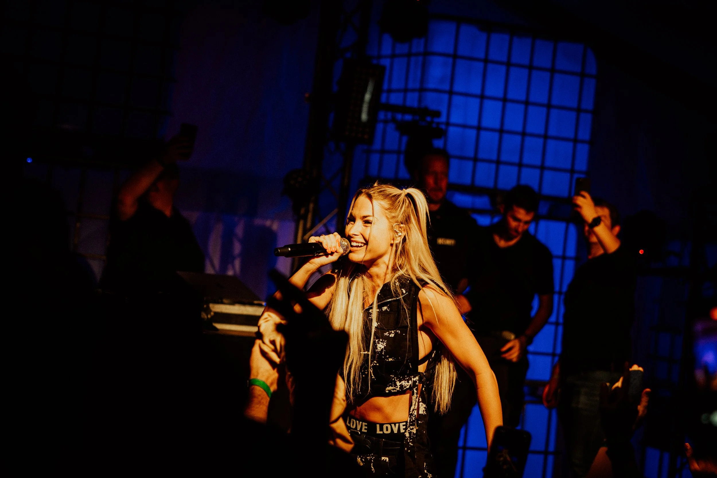 Junge Sängerin mit langen blonden Haaren, singt in Mikrofon bei Konzert, im Hintergrund sind Musiker und Bühnenbeleuchtung, blauer Hintergrundlichteffekt.