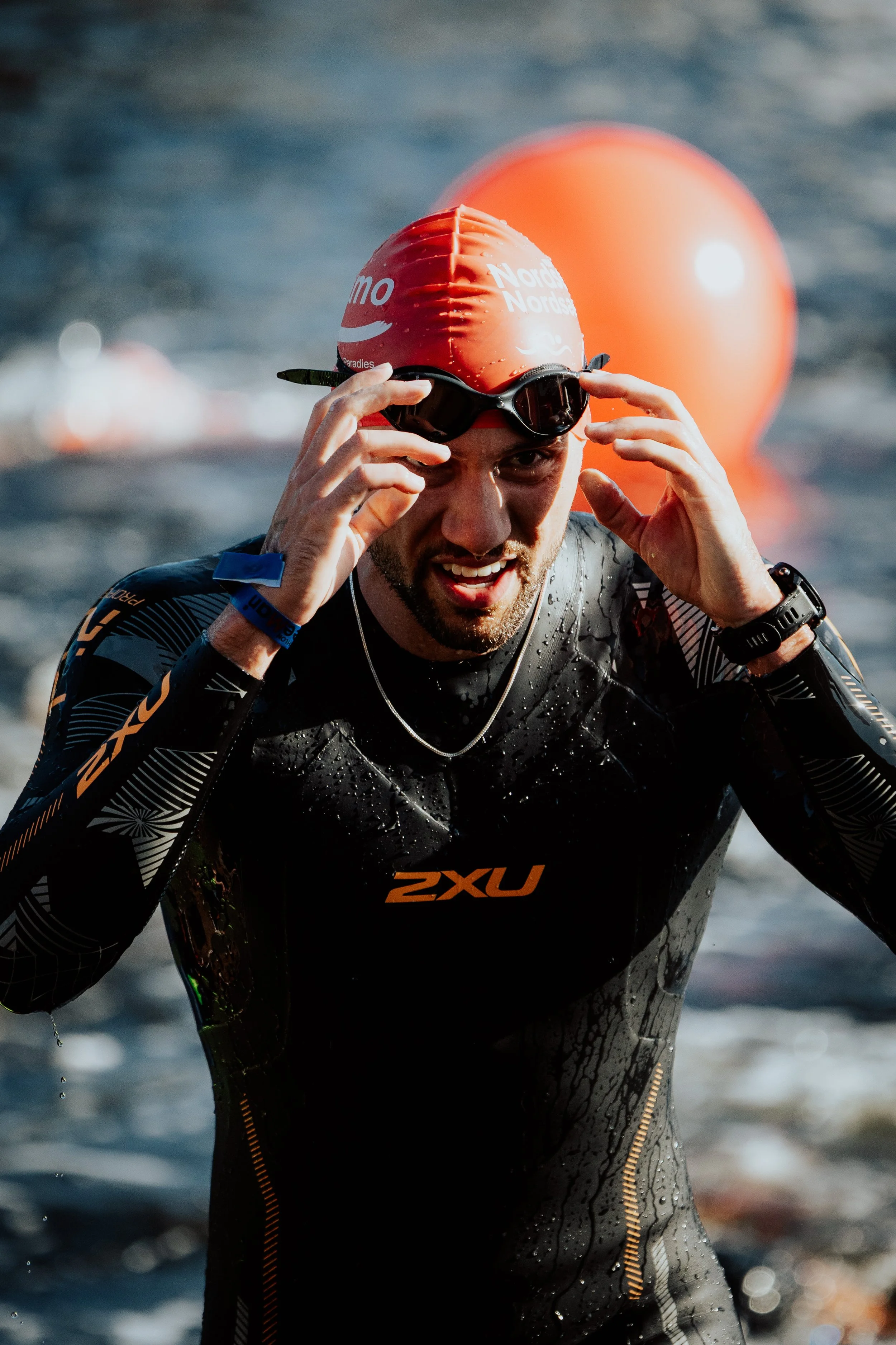 Schwimmer trägt rote Badekappe und taucherbrille, steht im Wasser, trägt einen schwarzen Neoprenanzug mit orangefarbenen Akzenten, im Hintergrund Wasser und eine orange Boje.