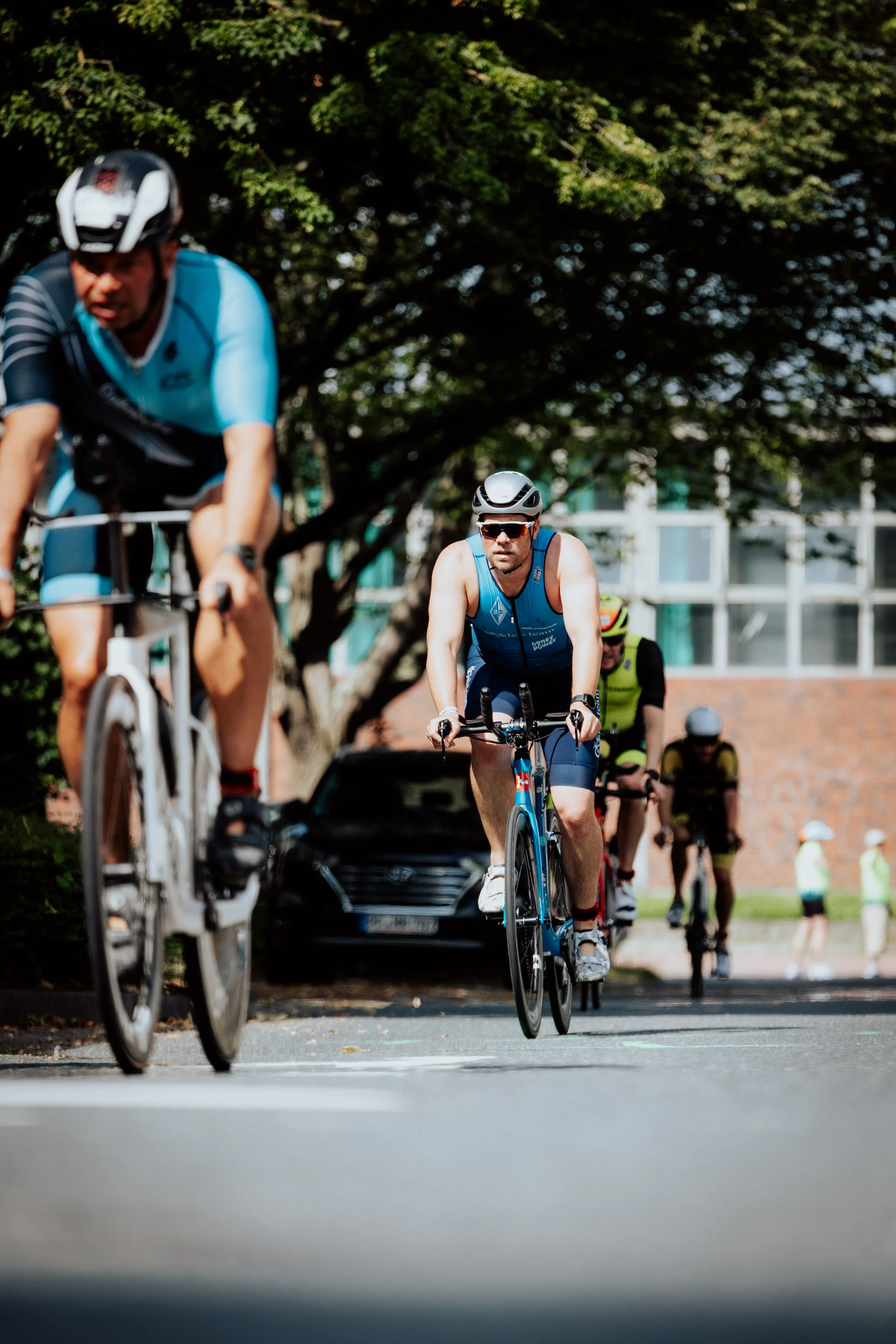 Mehrere Radfahrer beim Rennen auf einer Stadtstraße, mit Bäumen und Gebäuden im Hintergrund, Blick auf das Fahrrad eines Teilnehmers im Vordergrund.