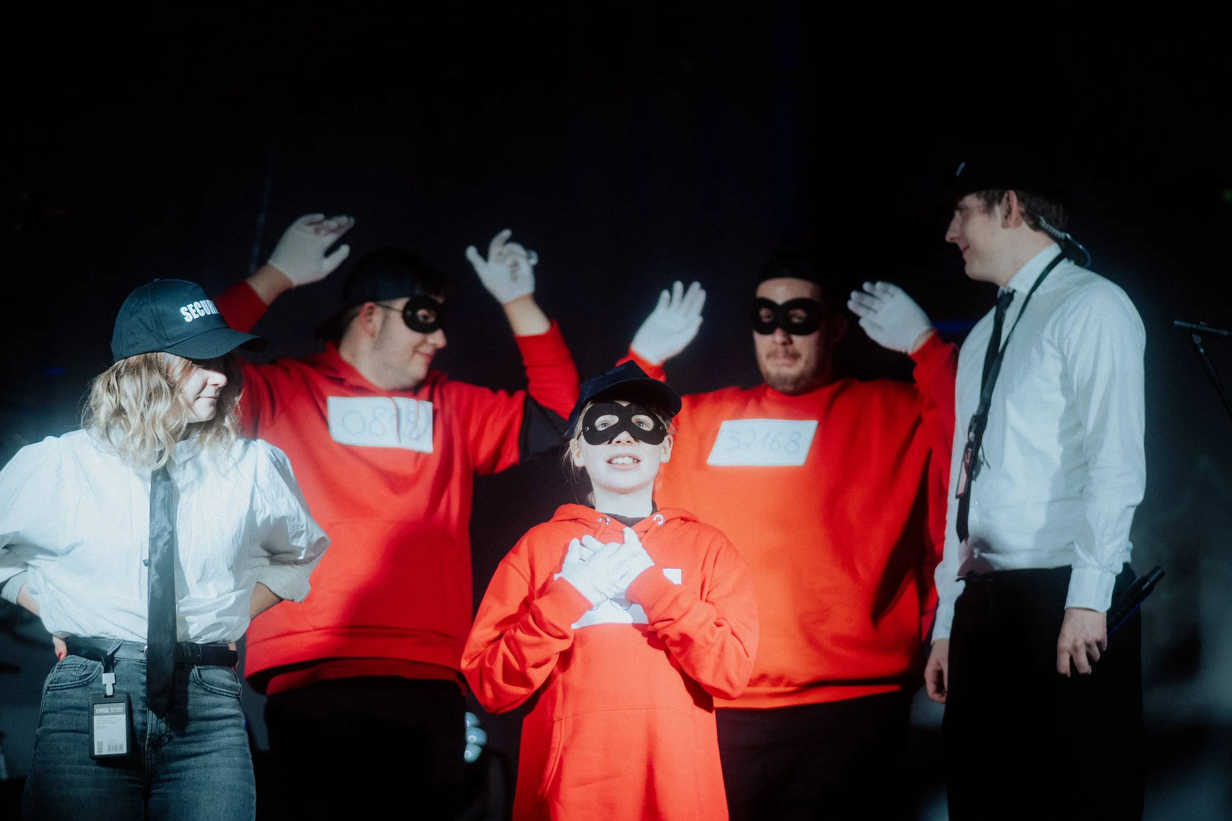 Menschen, die an einem Rollenspiel oder Theaterstück teilnehmen, mit einer Person in einem weißen Hemd, die als Regisseur oder Leiter hat, und mehreren Personen in roten Kostümen mit schwarzen Masken, die sich für das Szenario vorbereiten.