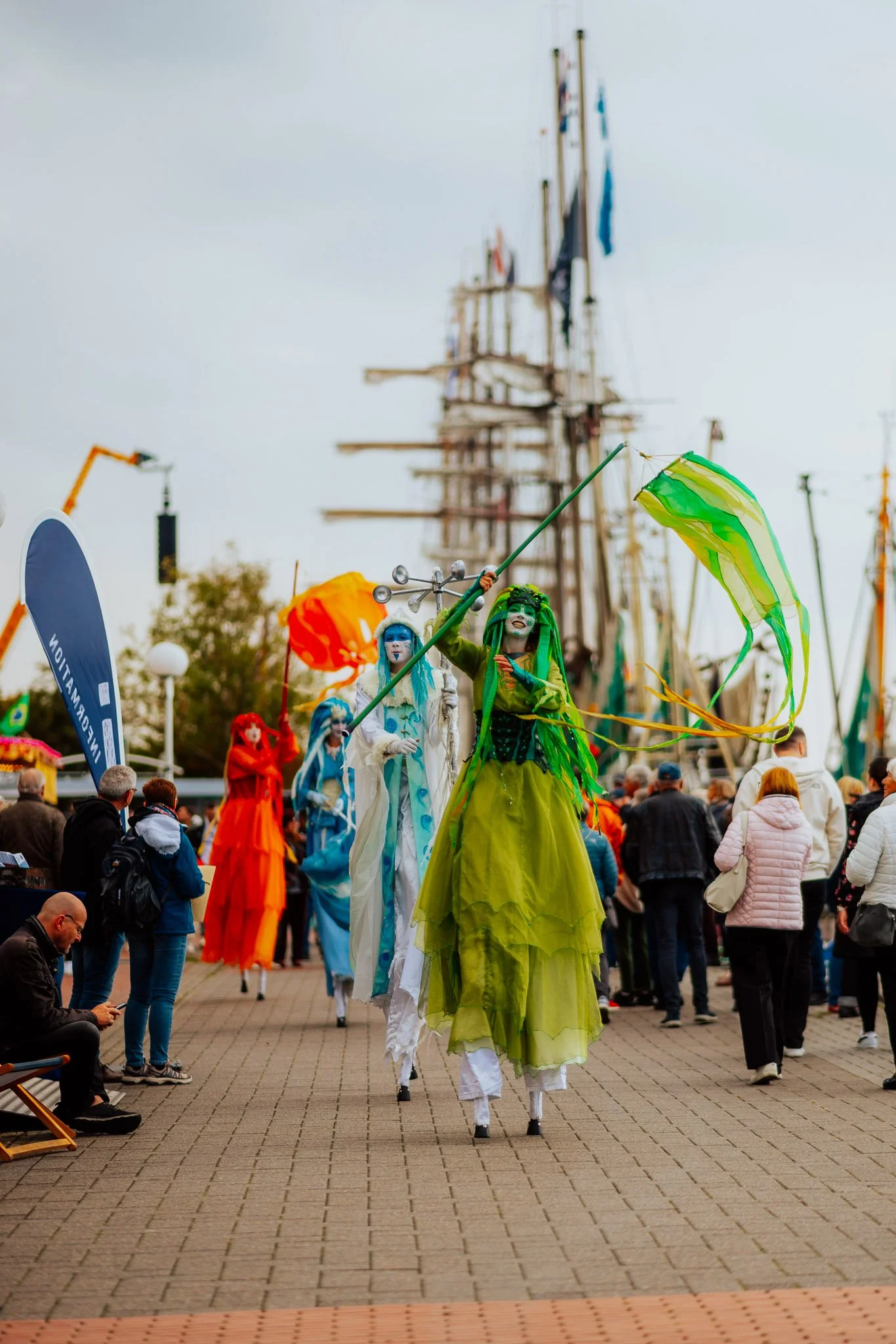 Mehrere große, bühnenartige Figuren in bunten Kostümen, die auf Stelzen laufen, in einer Stadtmitte während eines Festes oder Festivals. Im Hintergrund stehen Menschen, einige fotografieren oder schauen zu, und es gibt eine Turmstruktur, wahrscheinli