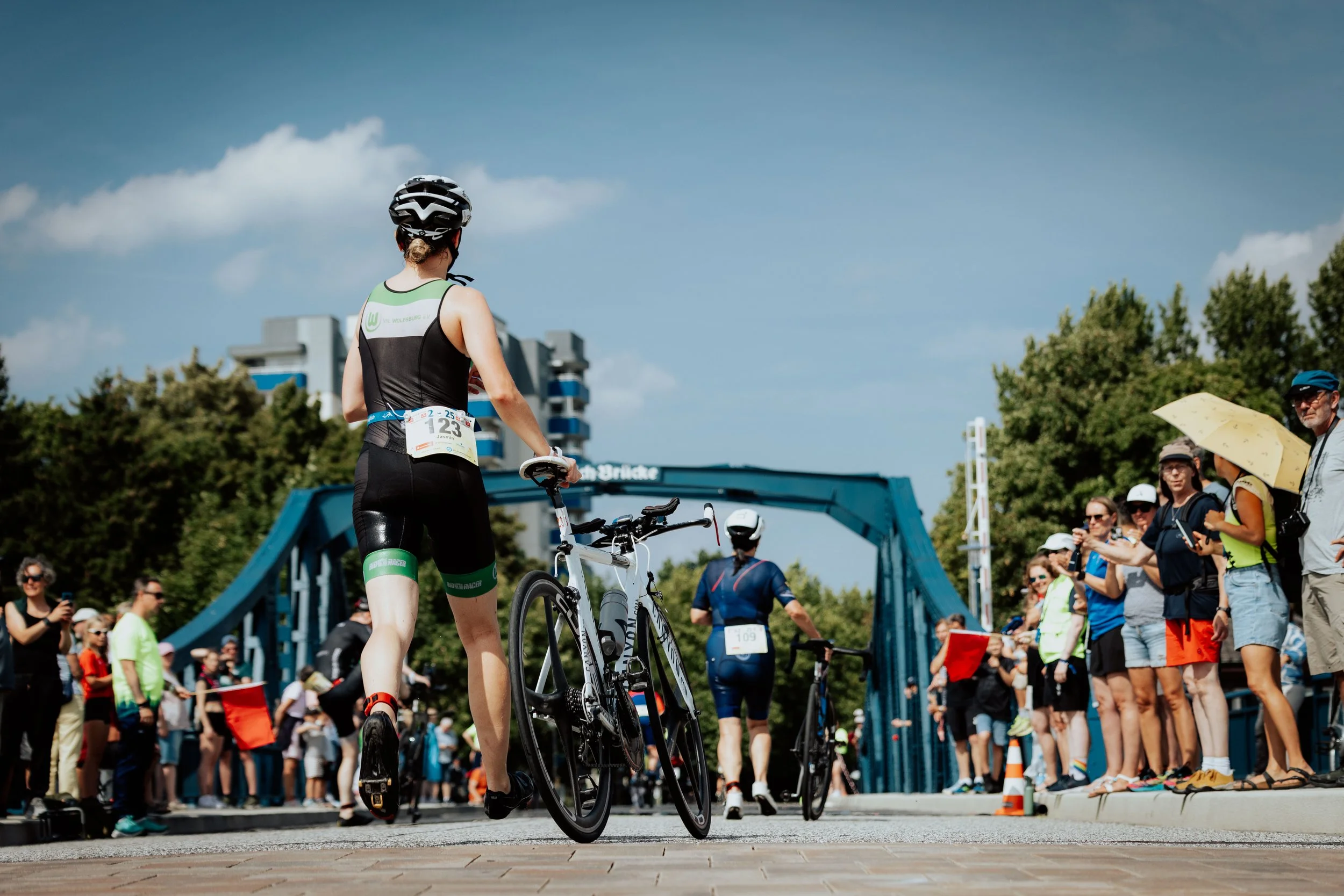 Eine Frau mit Fahrrad bei einem Triathlon-Event, umgeben von Zuschauern und Veranstaltungspersonal, im Freien bei sonnigem Wetter.