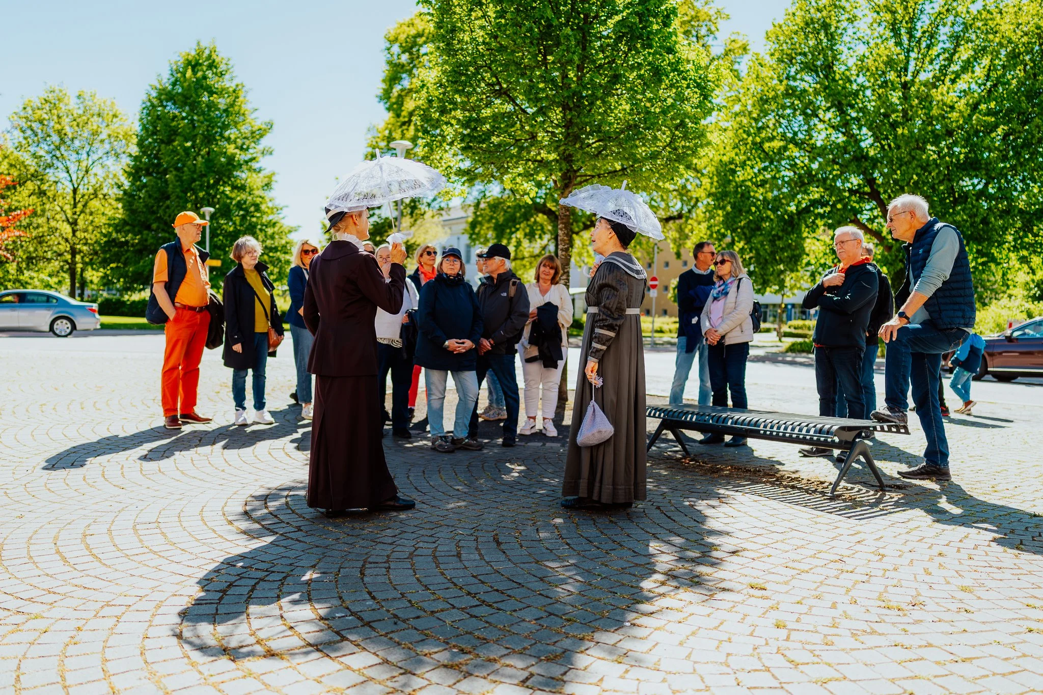 Gruppe von älteren Menschen, die im Park stehen, während zwei Damen mit altenoriösen Kleidung und Sonnenparasols sprechen. Es scheint eine Tour oder Diskussion im Freien zu sein, bei sonnigem Wetter, umgeben von grünen Bäumen.