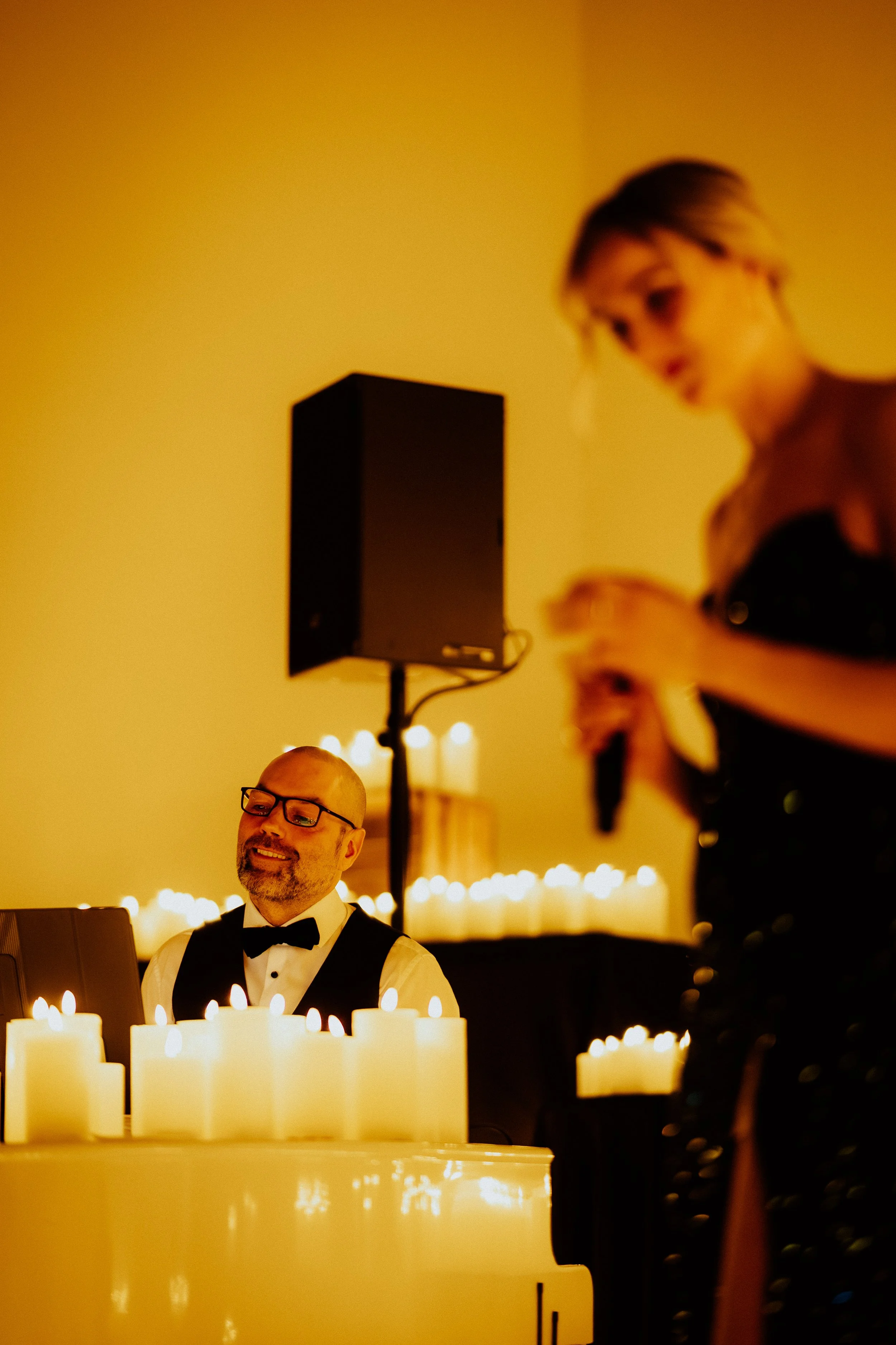 Ein Mann in Smoking mit Krawatte spielt Klavier in einem festlich beleuchteten Raum, umgeben von Kerzen.
