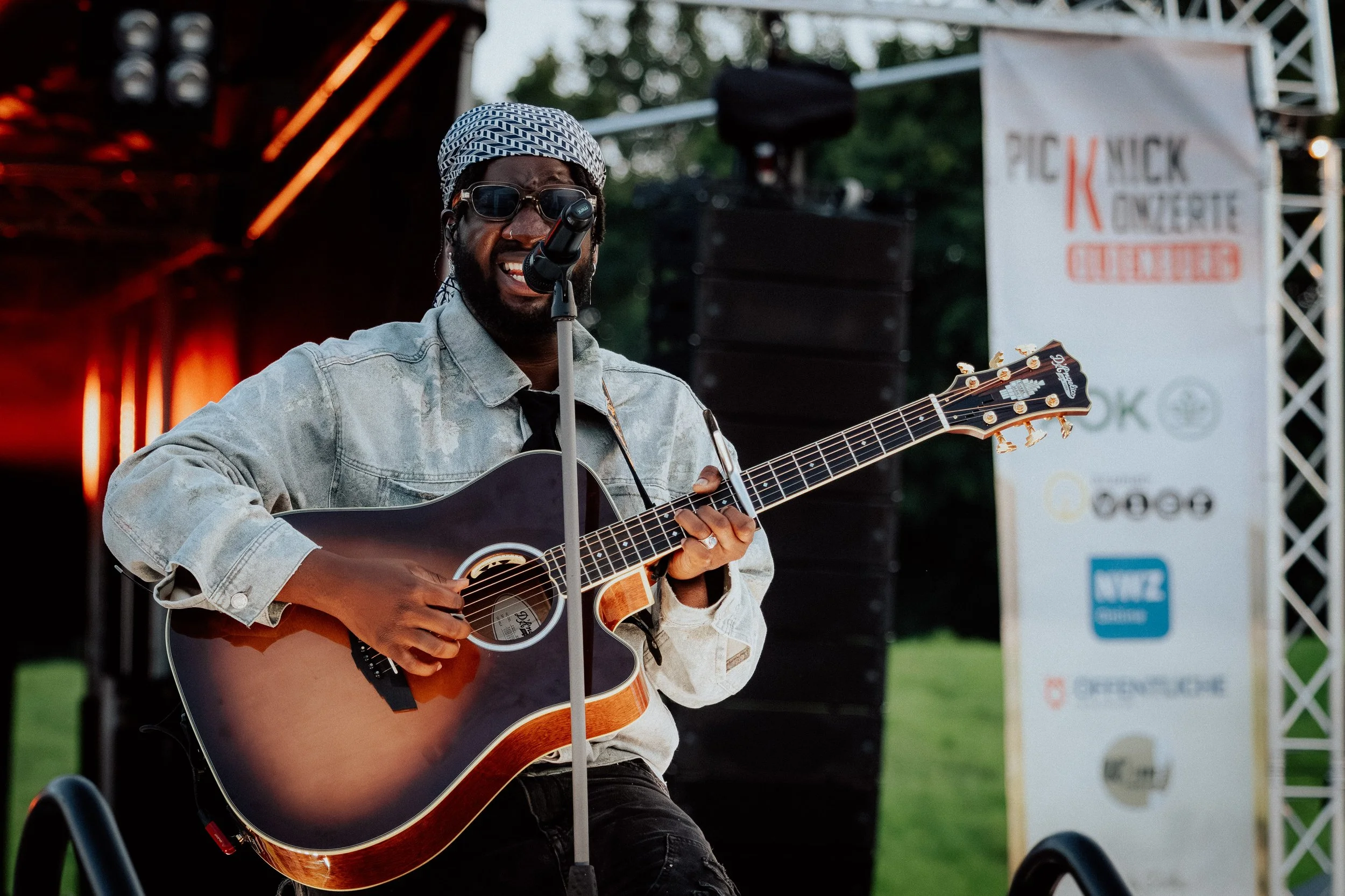 Mann singt und spielt Gitarre auf einer Bühne bei einem Open-Air-Konzert, im Hintergrund ein Banner mit dem Logo und Text. Der Mann trägt eine Mütze, Sonnenbrille und eine Jacke.
