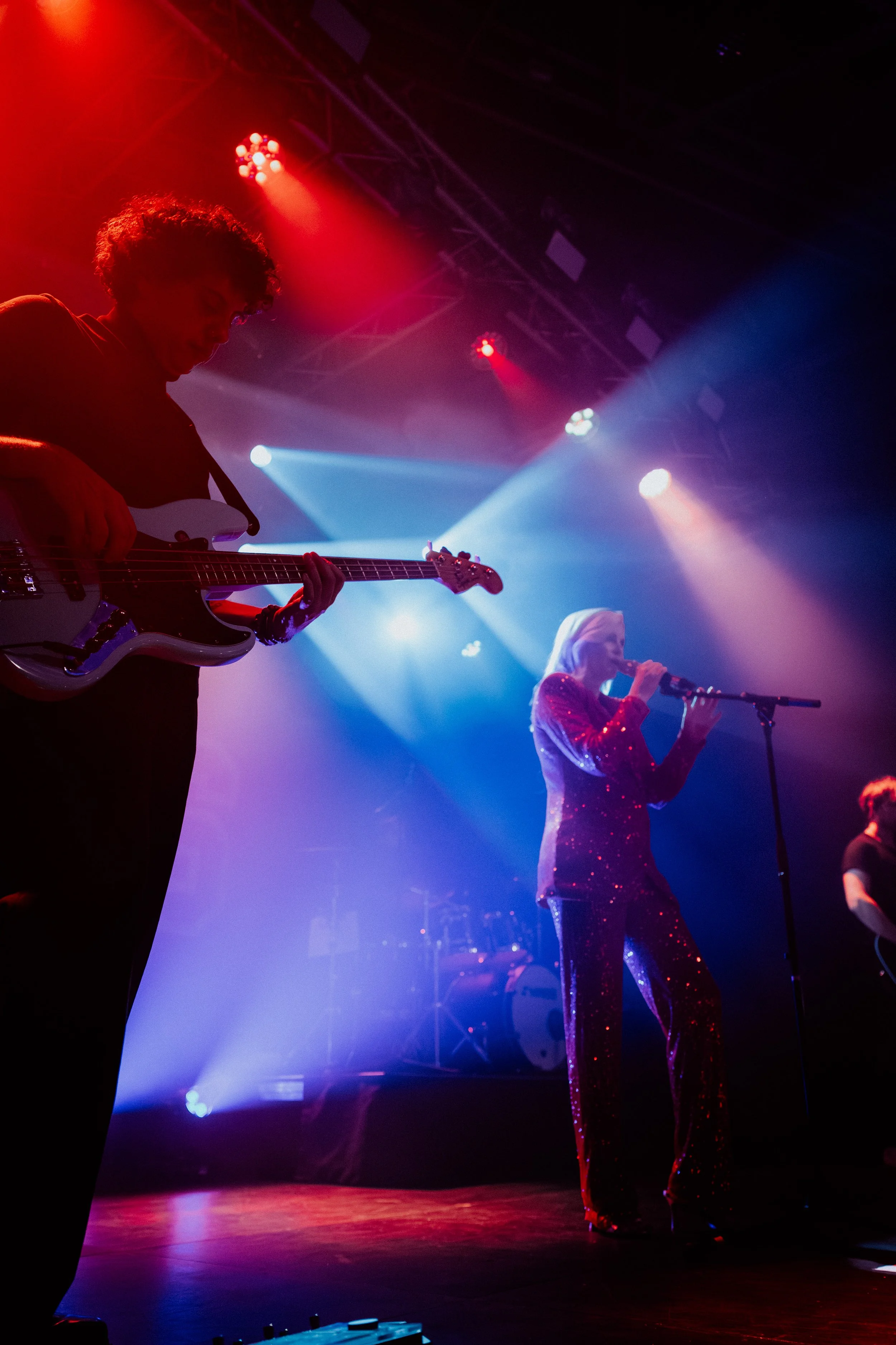 Musiker auf einer Bühne bei einem Konzert, mit buntem Bühnenlicht in Blau, Rot und Lila, ein Mann spielt E-Gitarre, eine Frau singt ins Mikrofon, beide sind in glamourösen Outfits gekleidet.