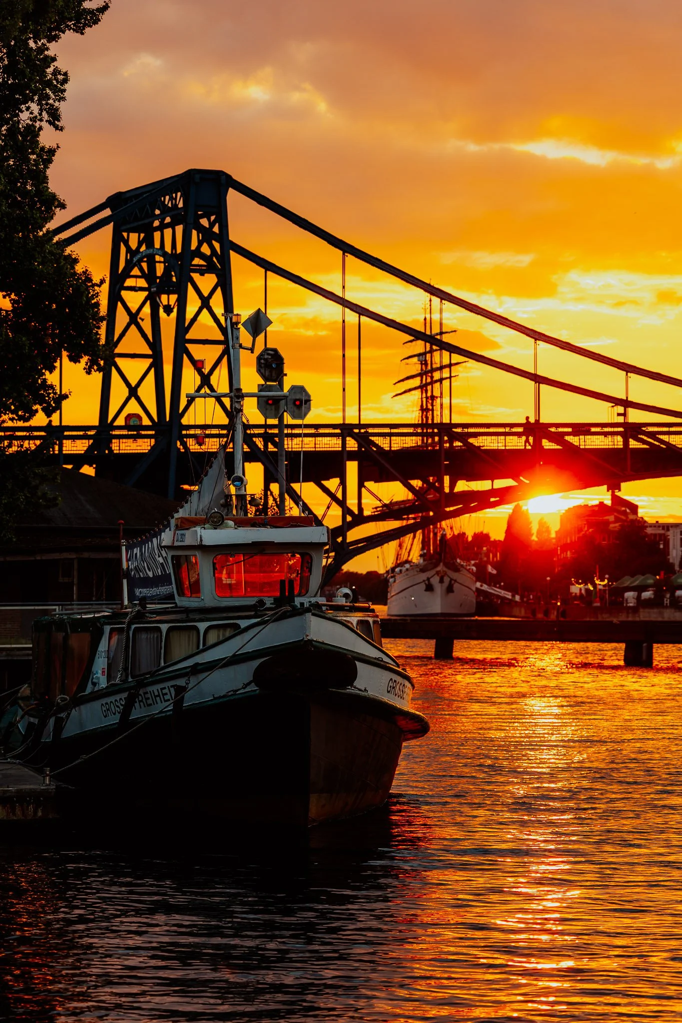 Ein Boot liegt im Wasser vor einer Brücke während eines Sonnenuntergangs, mit orangefarbenem Himmel im Hintergrund.
