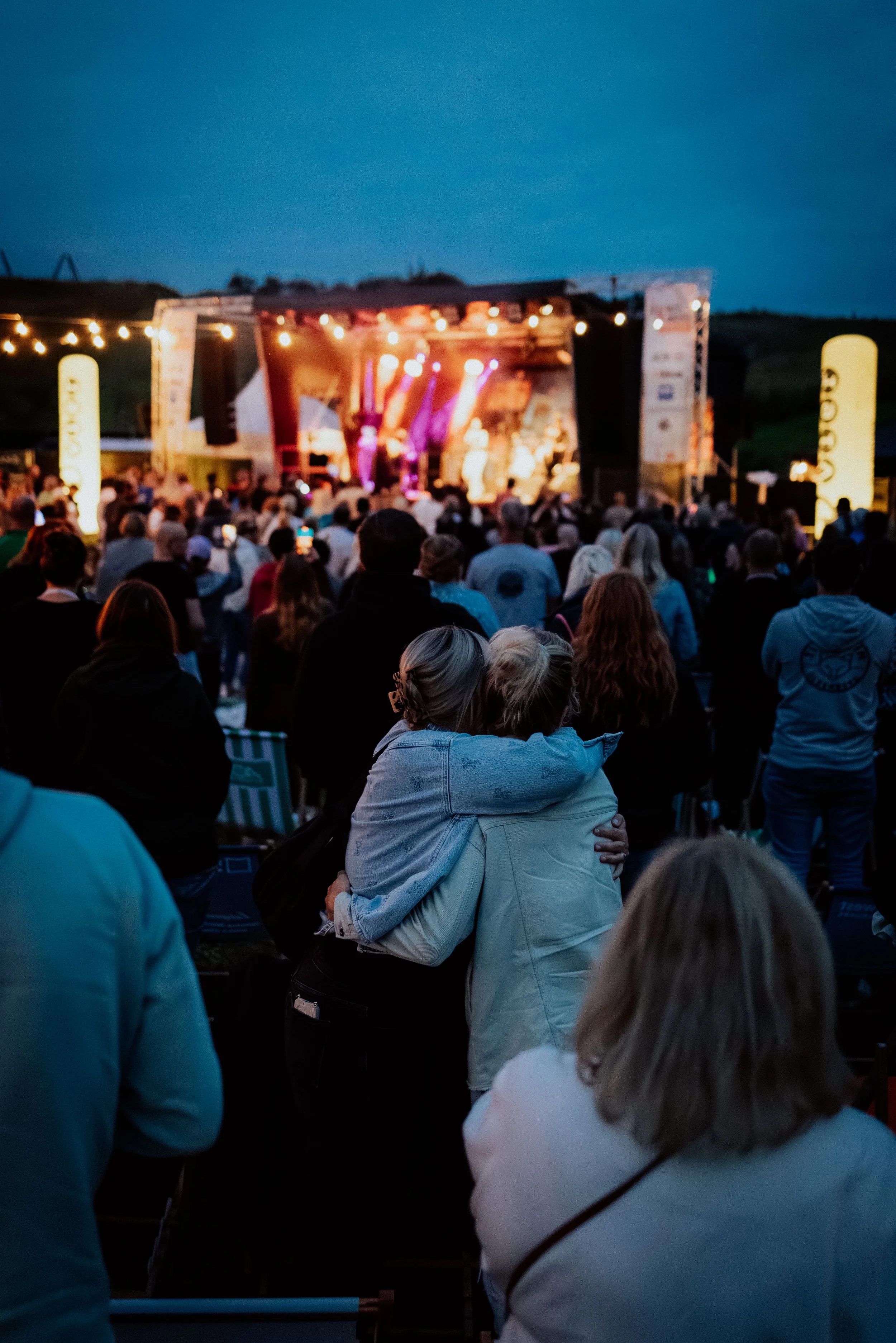 Menschen auf einem Konzert im Freien bei Nacht, im Hintergrund Bühne mit bunten Lichtern.
