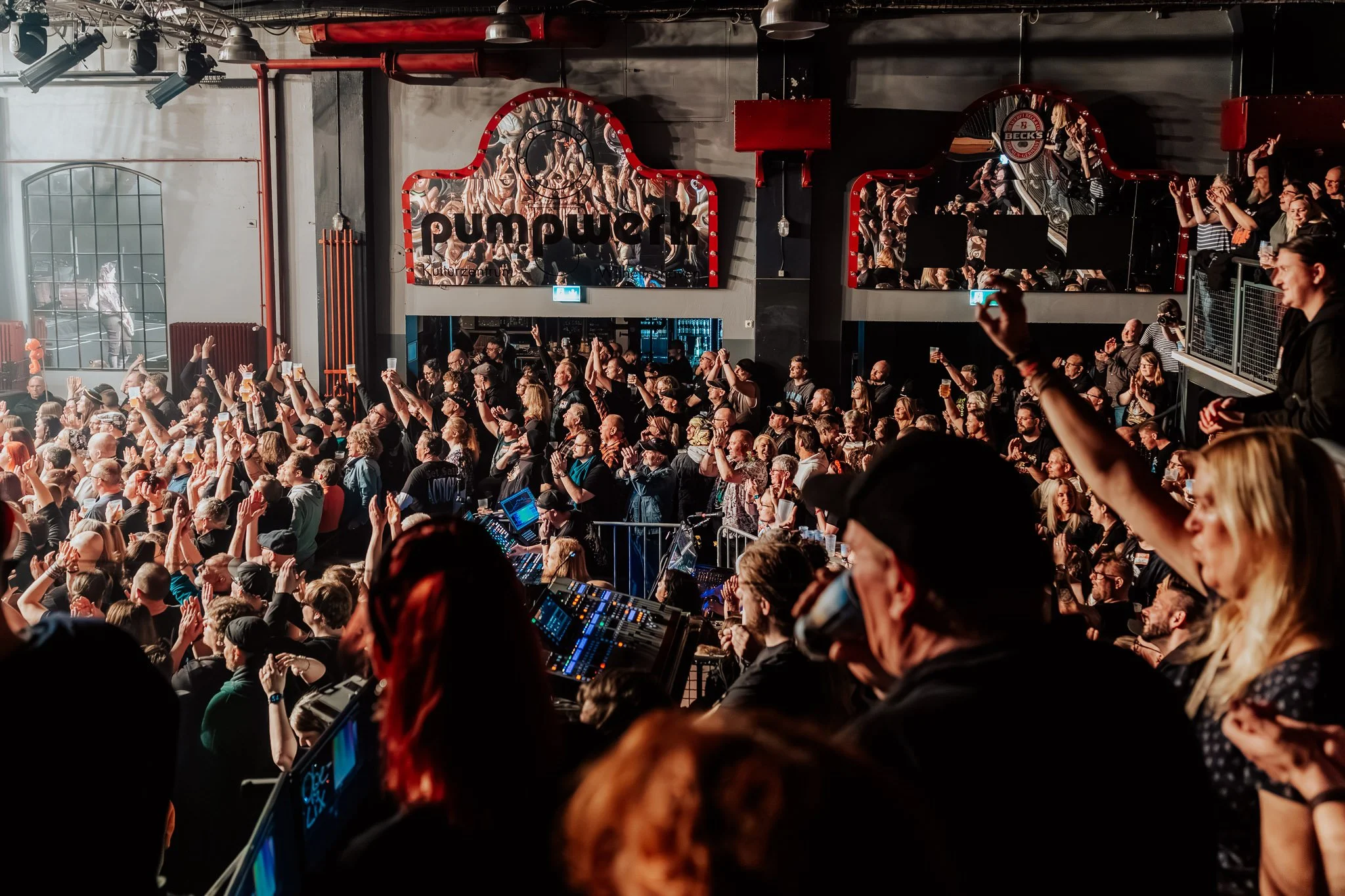 Innenaufnahme eines voll besetzten Konzert- oder Veranstaltungsortes mit Bühne, Publikum, Monitoren, Lichttechnik und einem großen Bildschirm mit der Aufschrift "pumpek".