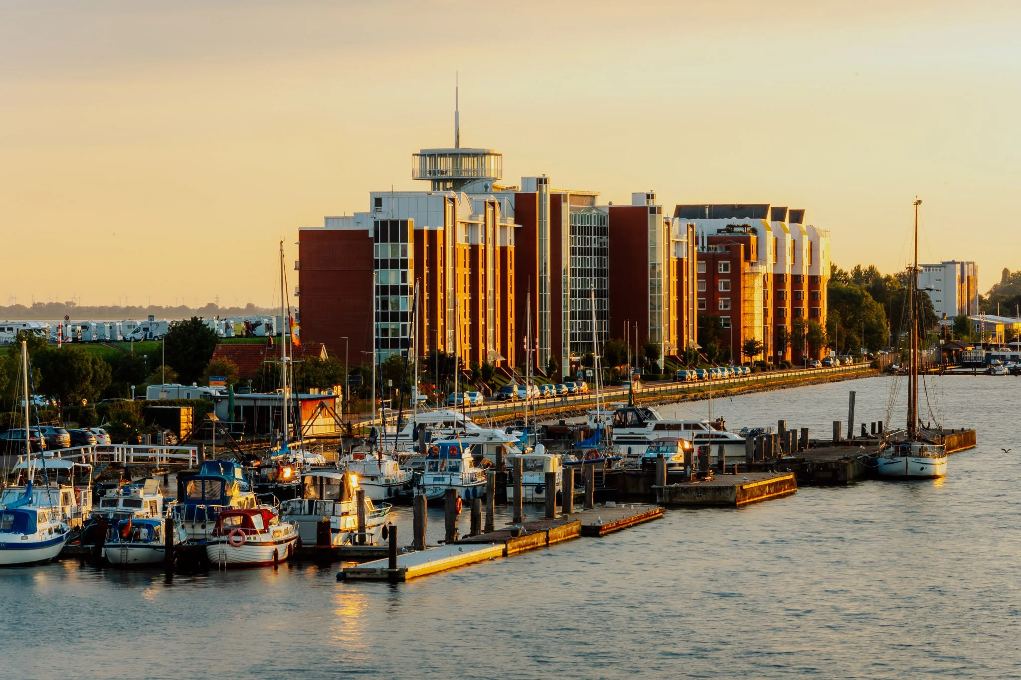 Blick auf einen Wasserhafen mit vielen Booten und Yachten bei Sonnenuntergang, im Hintergrund modern gestaltete Wohnhäuser am Wasser.