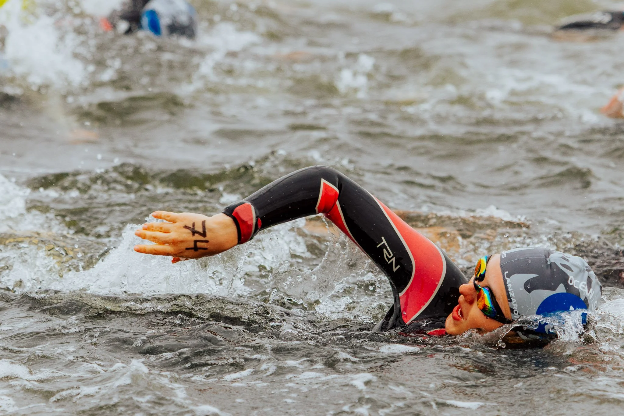 Ein Wassersportler beim Kraulschwimmen im natürlichen Fluss mit Gestein am Ufer, trägt eine Schwimmbrille und einen Neoprenanzug.