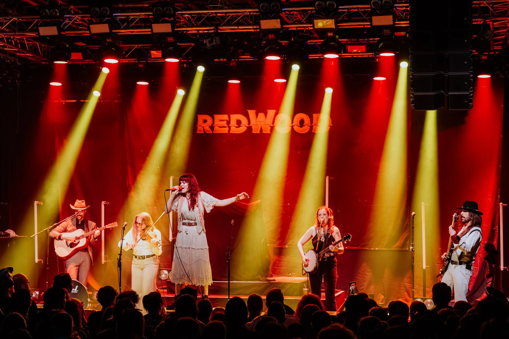 Auf einer Bühne performen mehrere Musiker und Sänger vor einem Publikum, mit rotem und gelbem Bühnenlicht, und einem roten Neon-Schriftzug, der "REDWOOD" lautet.