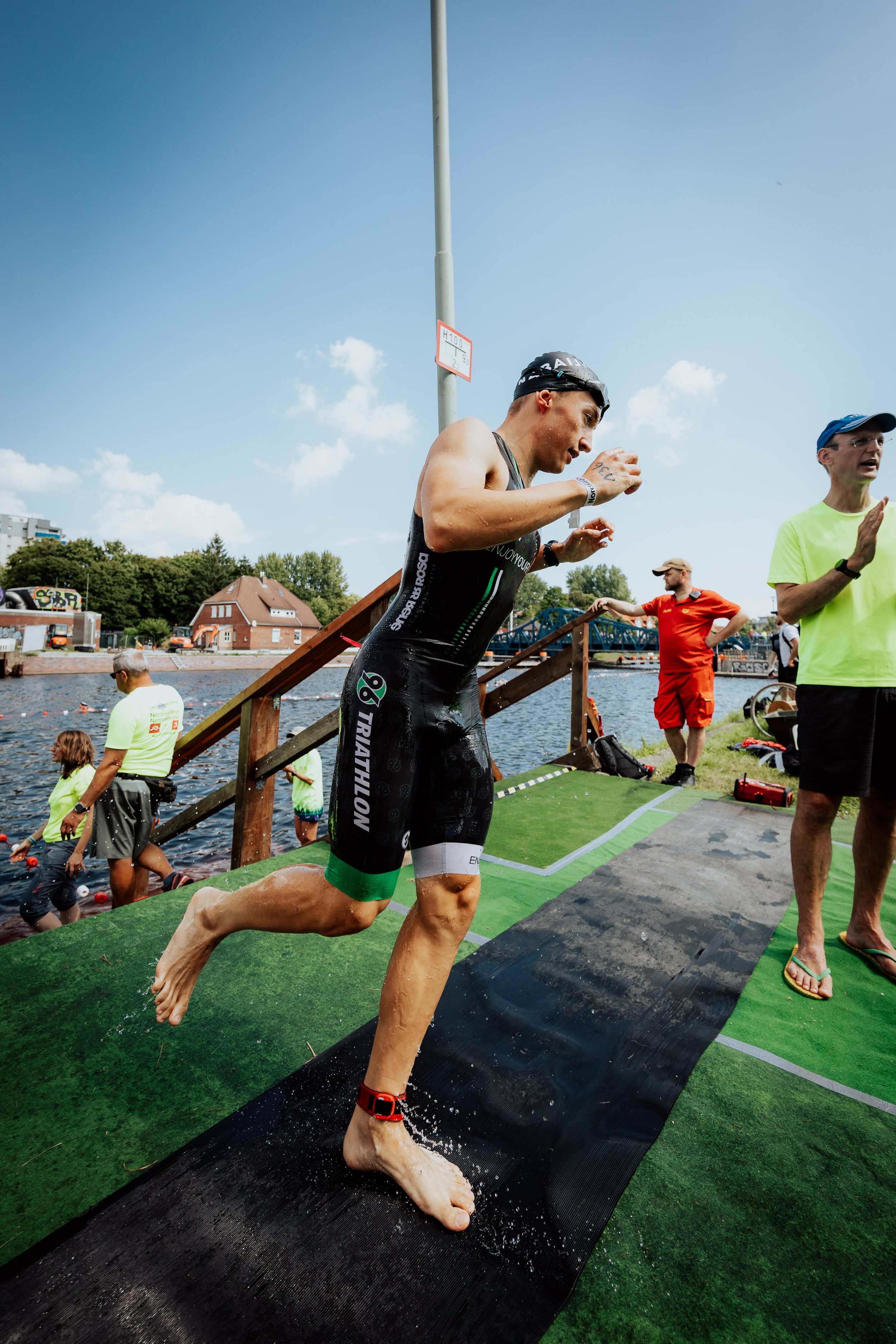Ein männlicher Triathlet beim Schwimmen, er steigt aus dem Wasser auf eine befestigte Plattform, um den Wettkampf fortzusetzen. Er trägt einen schwarzen Triathlonanzug, eine schwarze Badekappe und eine Armbanduhr, während andere Teilnehmer und Beglei