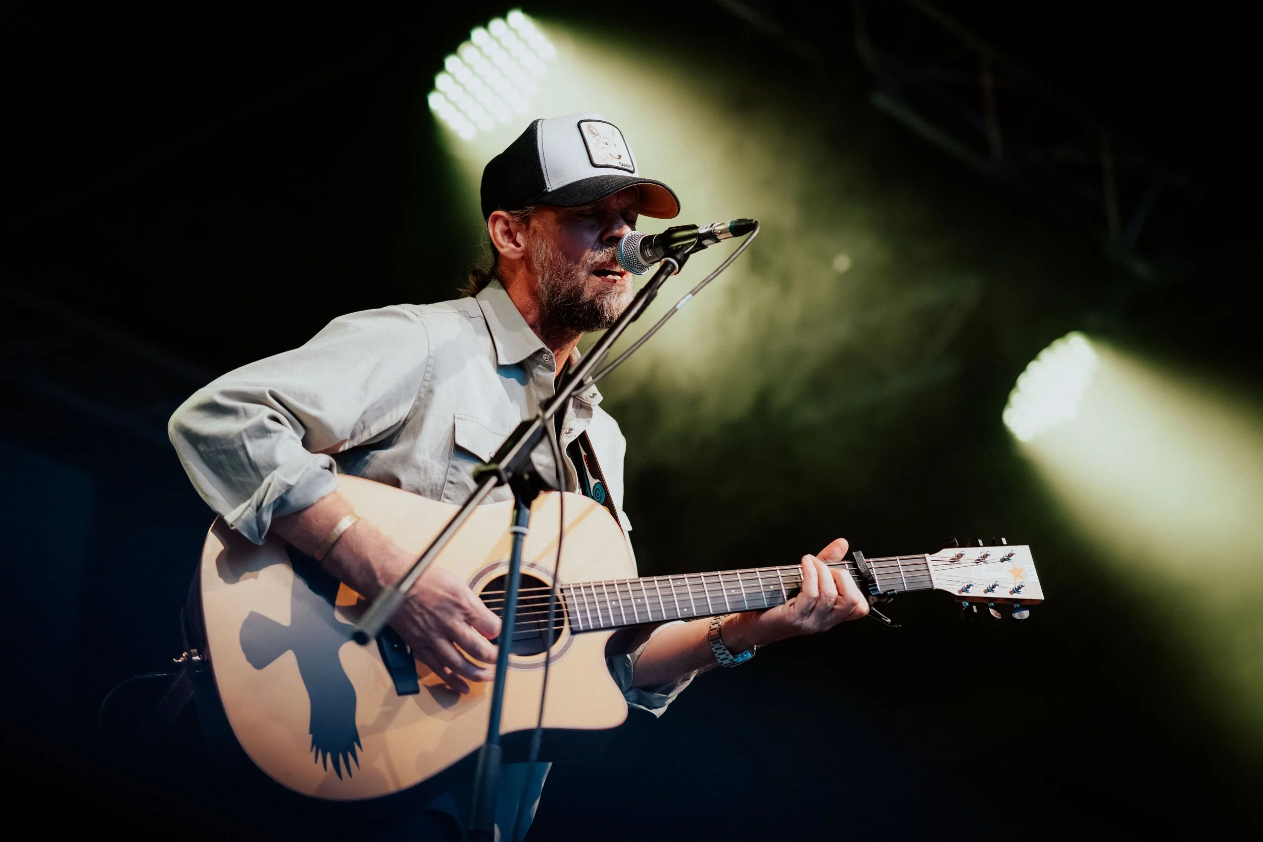 Mann spielt Gitarre und singt auf einer Bühne bei einem Konzert, hinter ihm sind Lichter und eine dunkle Kulisse.