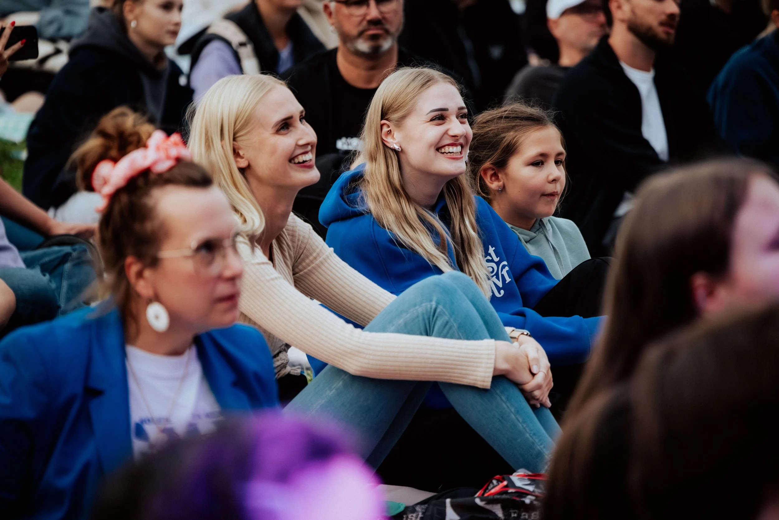 Menschen sitzen im Publikum bei einer Veranstaltung, lächeln und lachen, einige tragen Hoodies und T-Shirts, einige haben Hüte oder Ohrringe, im Hintergrund sind weitere Personen sichtbar.