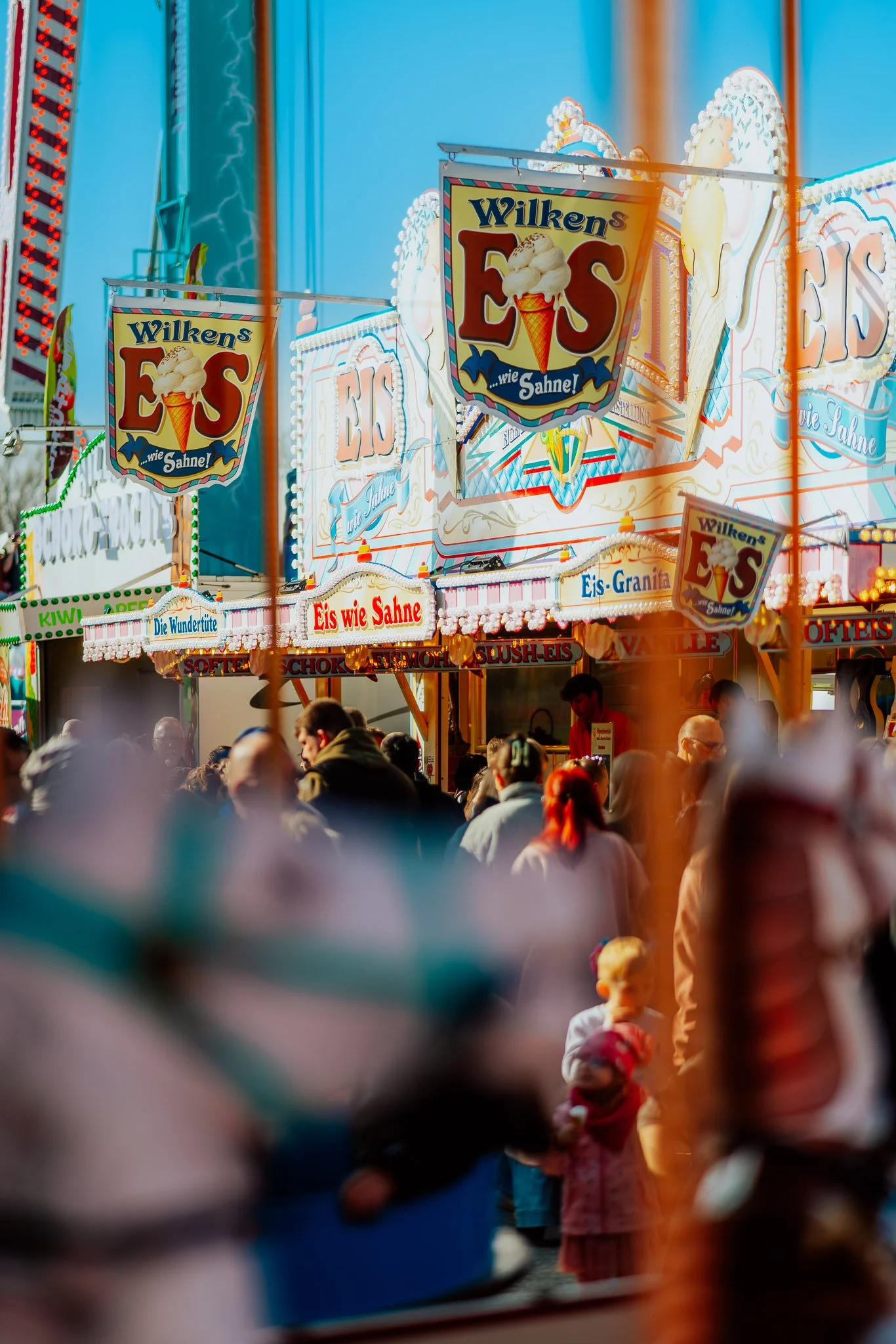Bunte Jahrmarktsszene mit Eisständen und Menschenmengen, im Hintergrund leuchtende Lichter und Schilder, fokussiert auf ein Eis-Stand mit dem Schild 'Wilkens Eis wie Sahne'.