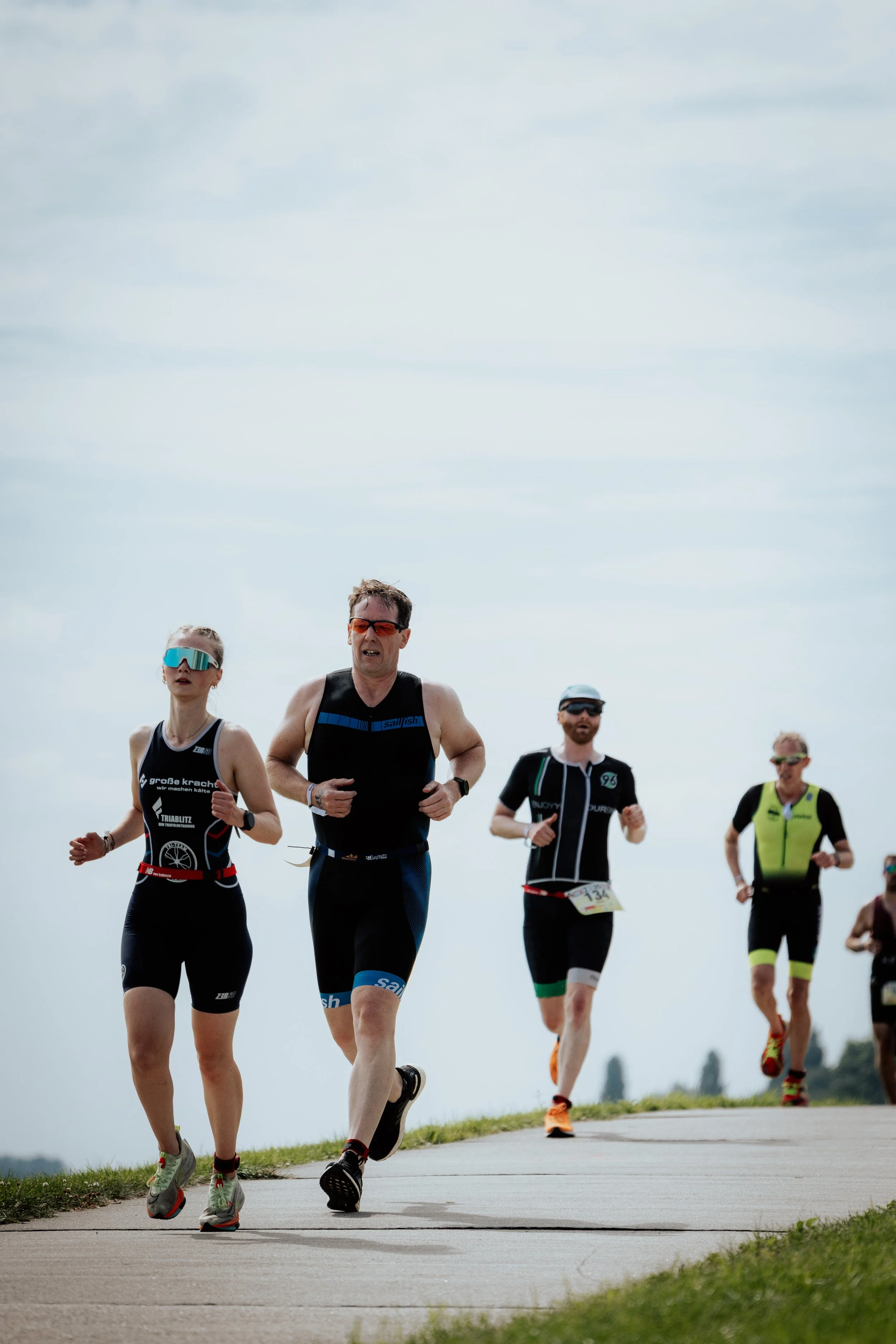 Mehrere Menschen laufen bei einem Marathonlauf auf einer Straße bei sonnigem Wetter. Manche tragen Sportbekleidung und Sonnenbrillen, während andere eine Startnummer am Körper haben.