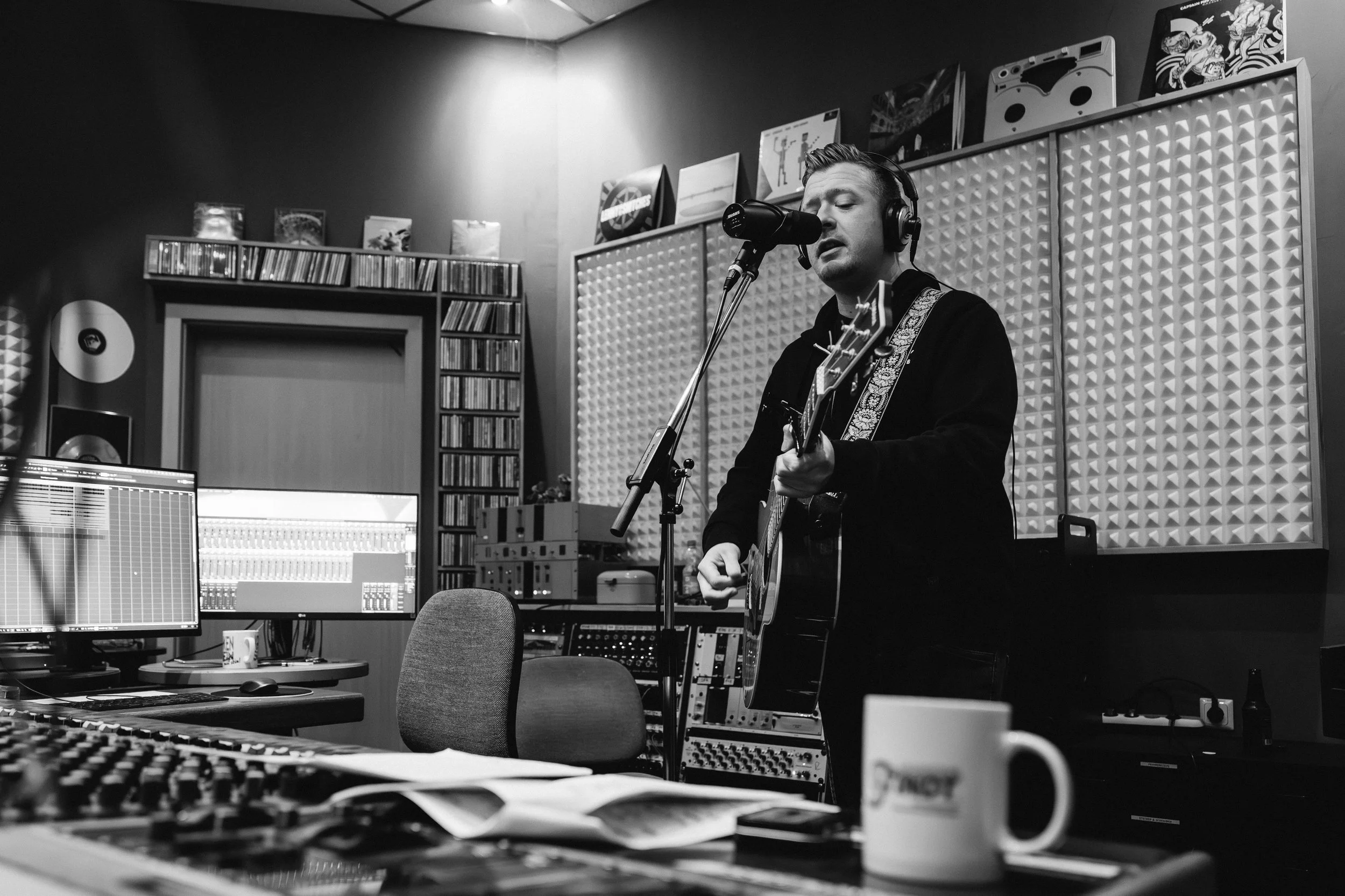 Ein Mann in einem Tonstudio spielt Gitarre und singt in einem Mikrofon, umgeben von Musikausrüstung und Hörerseln.
