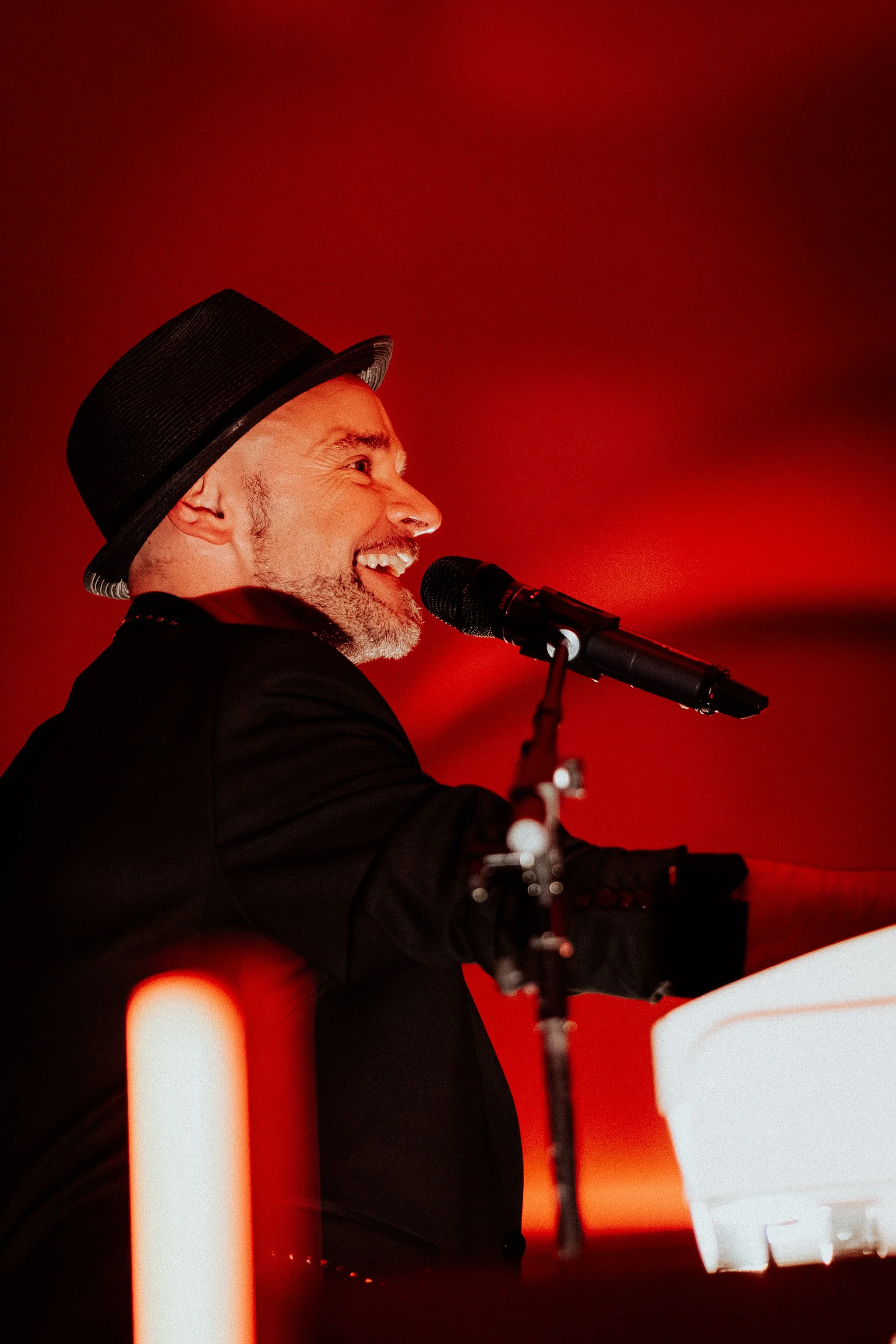 Ein Mann singt oder spricht in ein Mikrofon vor rotem Hintergrund, trägt einen schwarzen Anzug und einen schwarzen Hut.