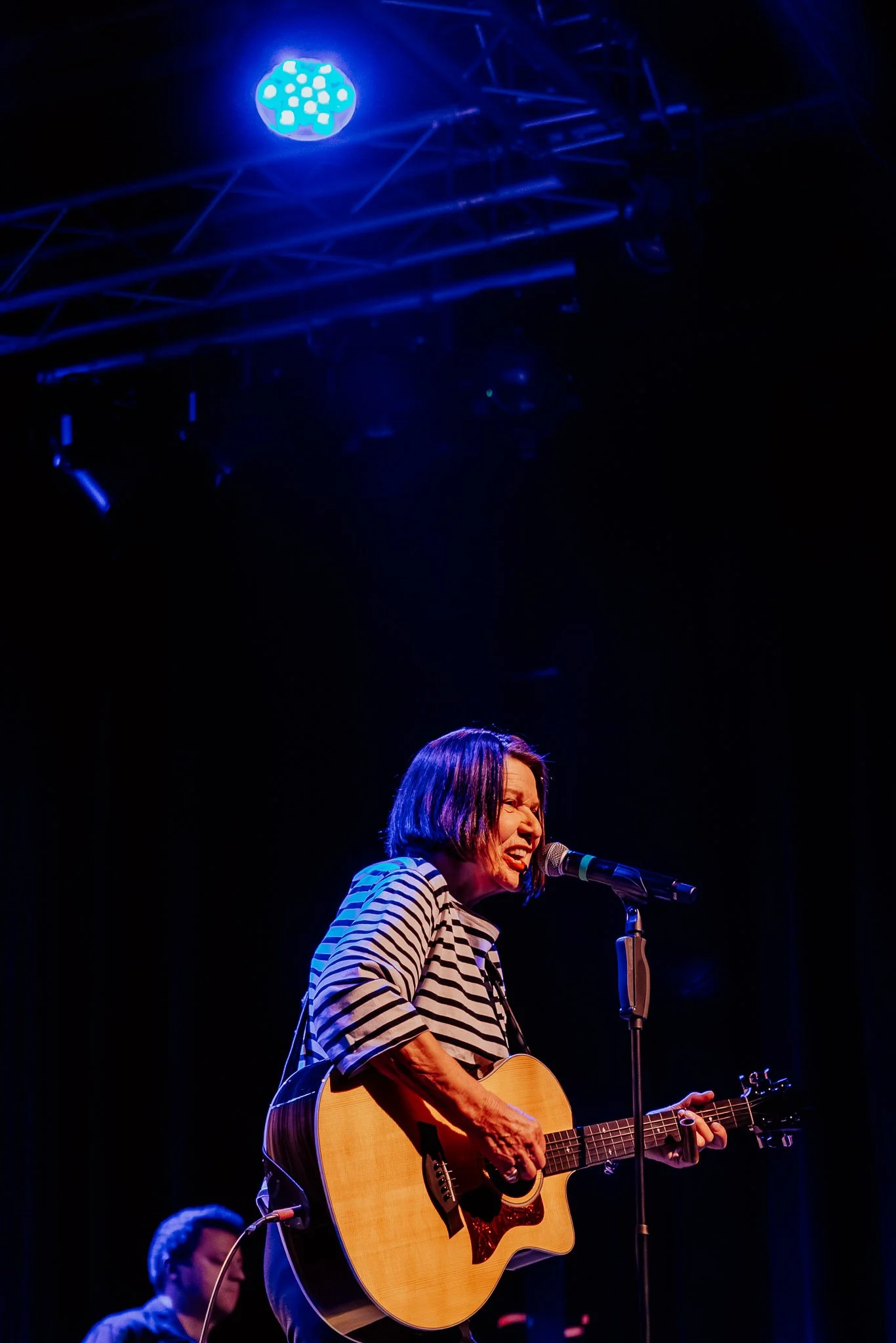 Eine Musikerin singt und spielt Gitarre auf einer Bühne, mit einem Mikrofon vor ihr, unter blauer Bühnenbeleuchtung.