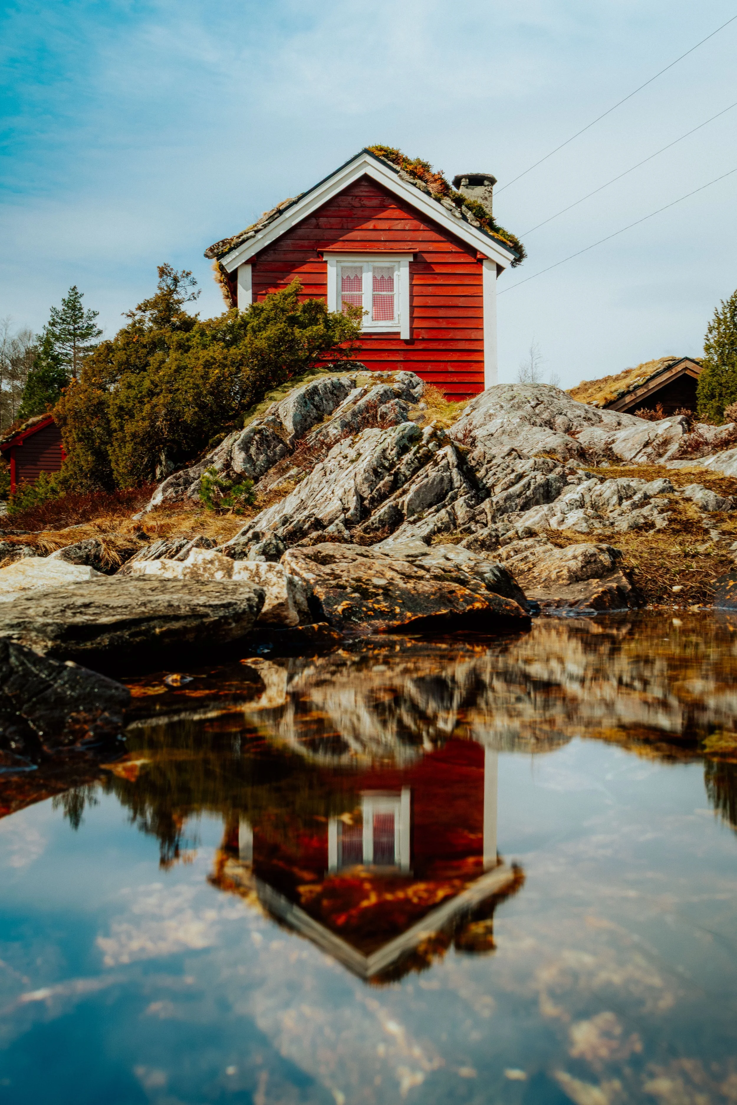 Ein rotes Holzhaus mit weißer Fensterrahmen und Schornstein, dessen Spiegelbild im Wasser eines Sees sichtbar ist, umgeben von Felsen und Bäumen bei sonnigem Himmel.