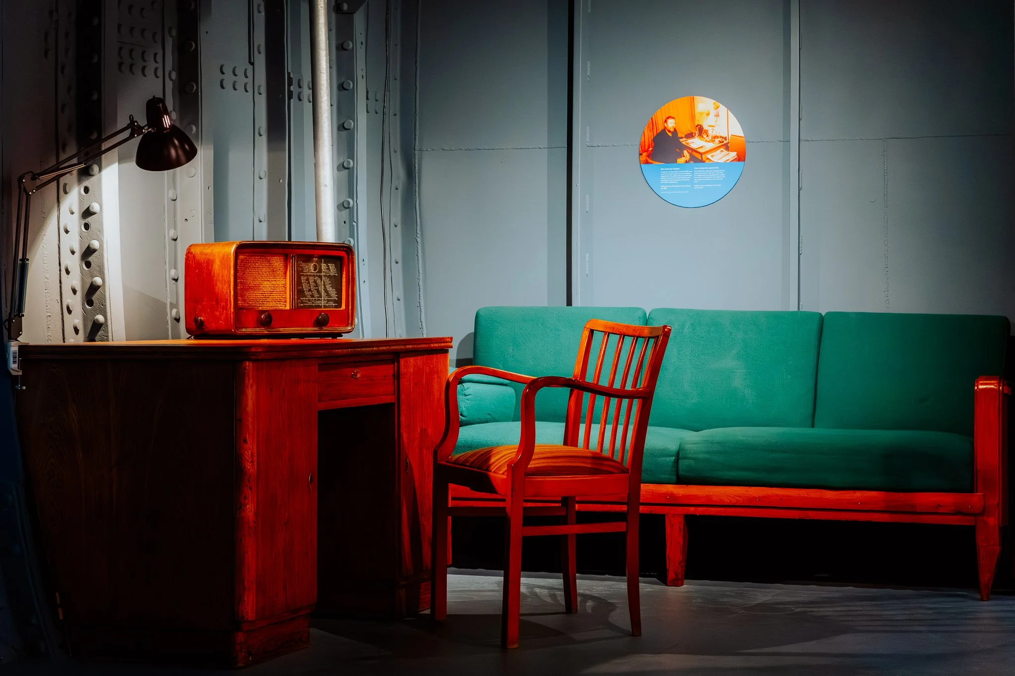 Ein Raum auf dem Museumsschiff Feuerschiff Norderney mit einem roten Radio, einem grünen Sofa und mehreren roten Stühlen, eine Lampe und eine Wandtafel