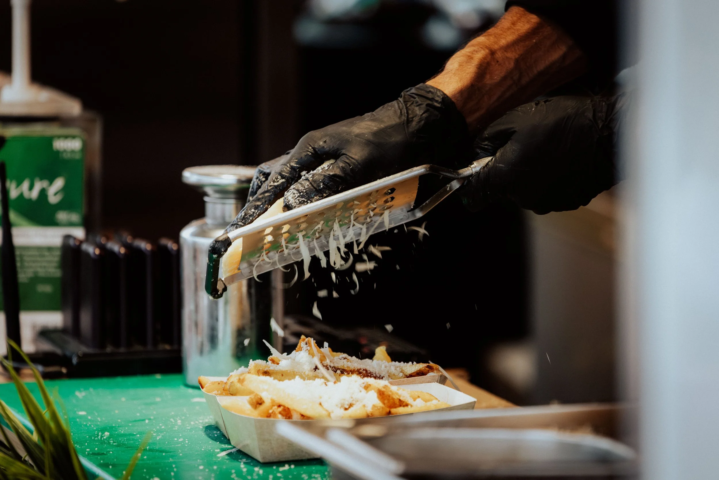 Eine Hand in einem schwarzen Handschuh reibt Käse auf eine Pasta. Darunter befindet sich eine weiße Schale mit Käse, die auf einer grünen Oberfläche steht.