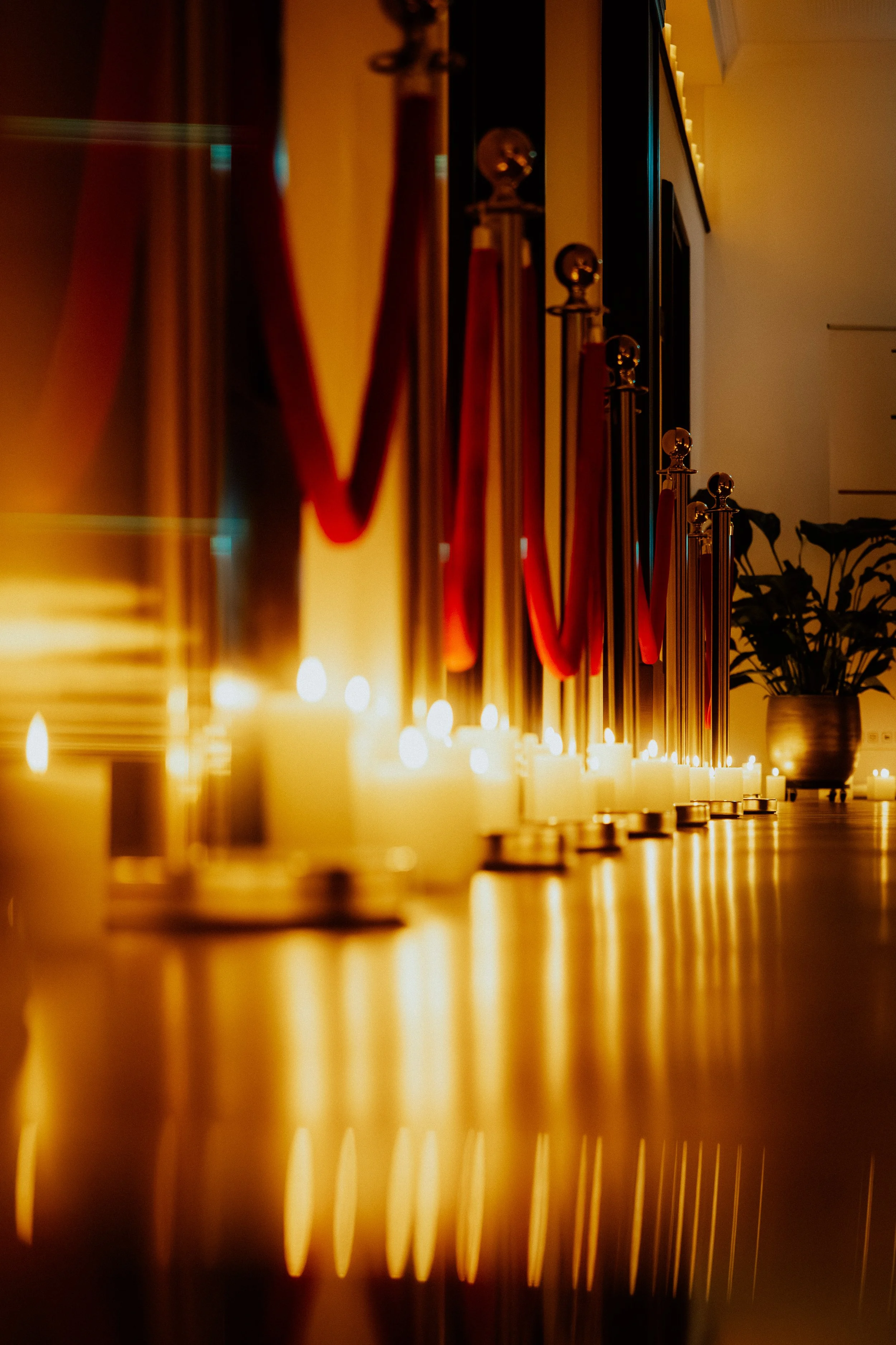 Eine Reihe von brennenden Kerzen auf einer Oberfläche, mit reflektierenden Metallpfosten im Hintergrund, und zugewandten Kerzen, mit warmem, gedämpftem Licht.