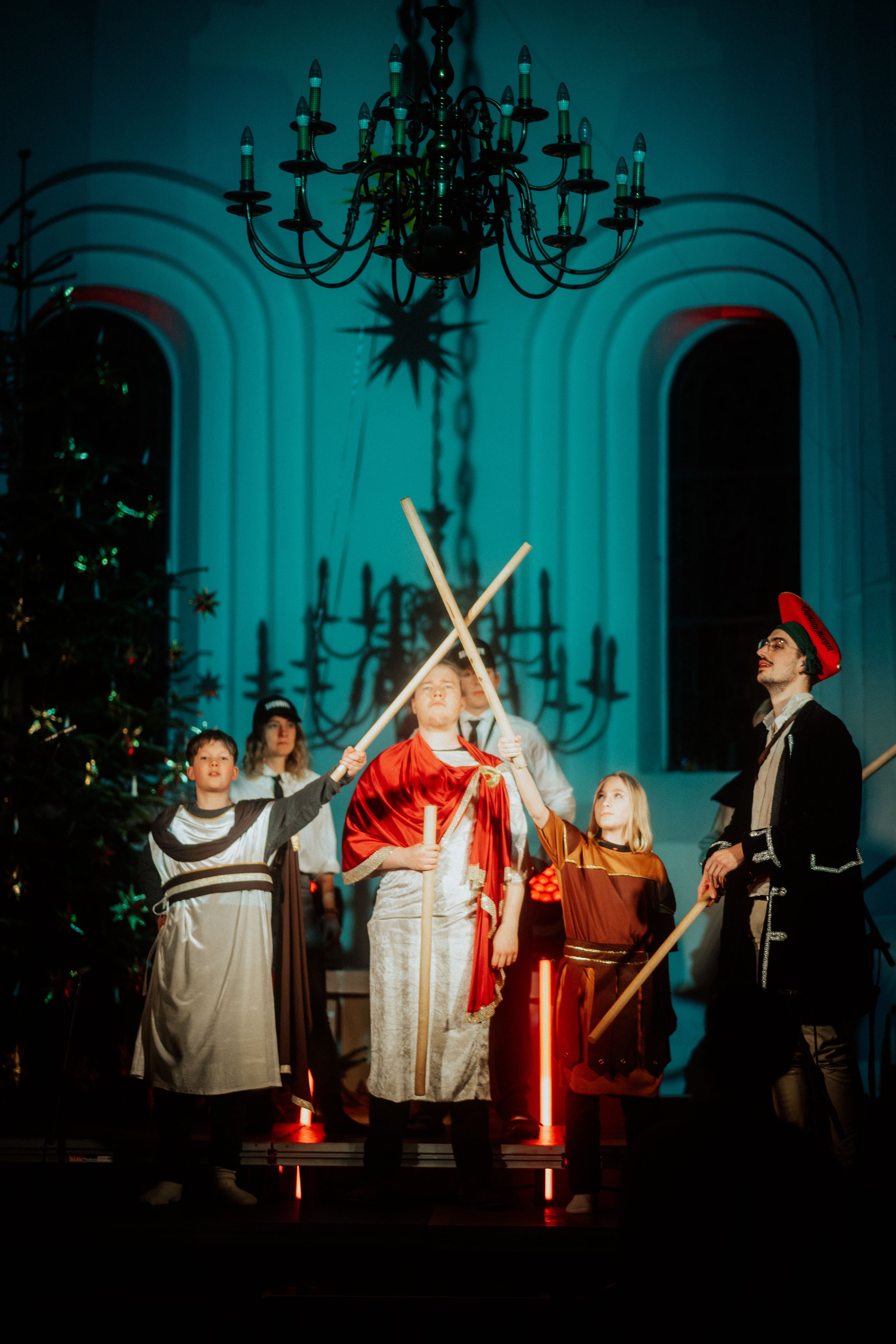 Sterne tragen Kostüme, die an biblische Figuren erinnern, bei einer Weihnachtsaufführung, mit Kerzenlichtern und einem Kronleuchter im Hintergrund.